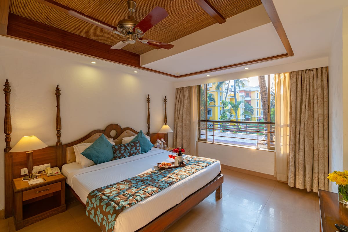 A hotel room with a large bed, wooden headboard, two bedside tables with lamps, a ceiling fan, and a window with curtains.