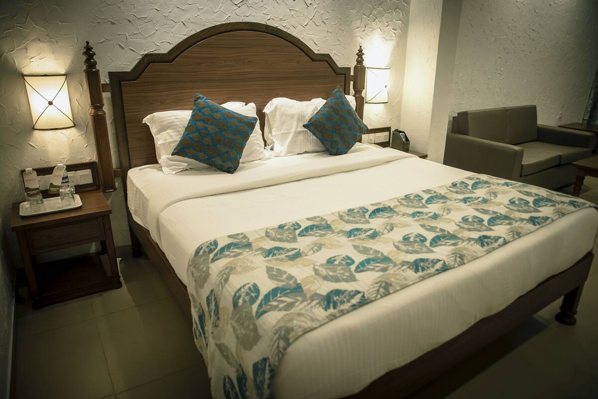 A neatly made bed with a floral bedspread, a wooden headboard, and two pillows with a geometric pattern. There is a nightstand with a lamp and a bottle of water. A chair is visible in the background.