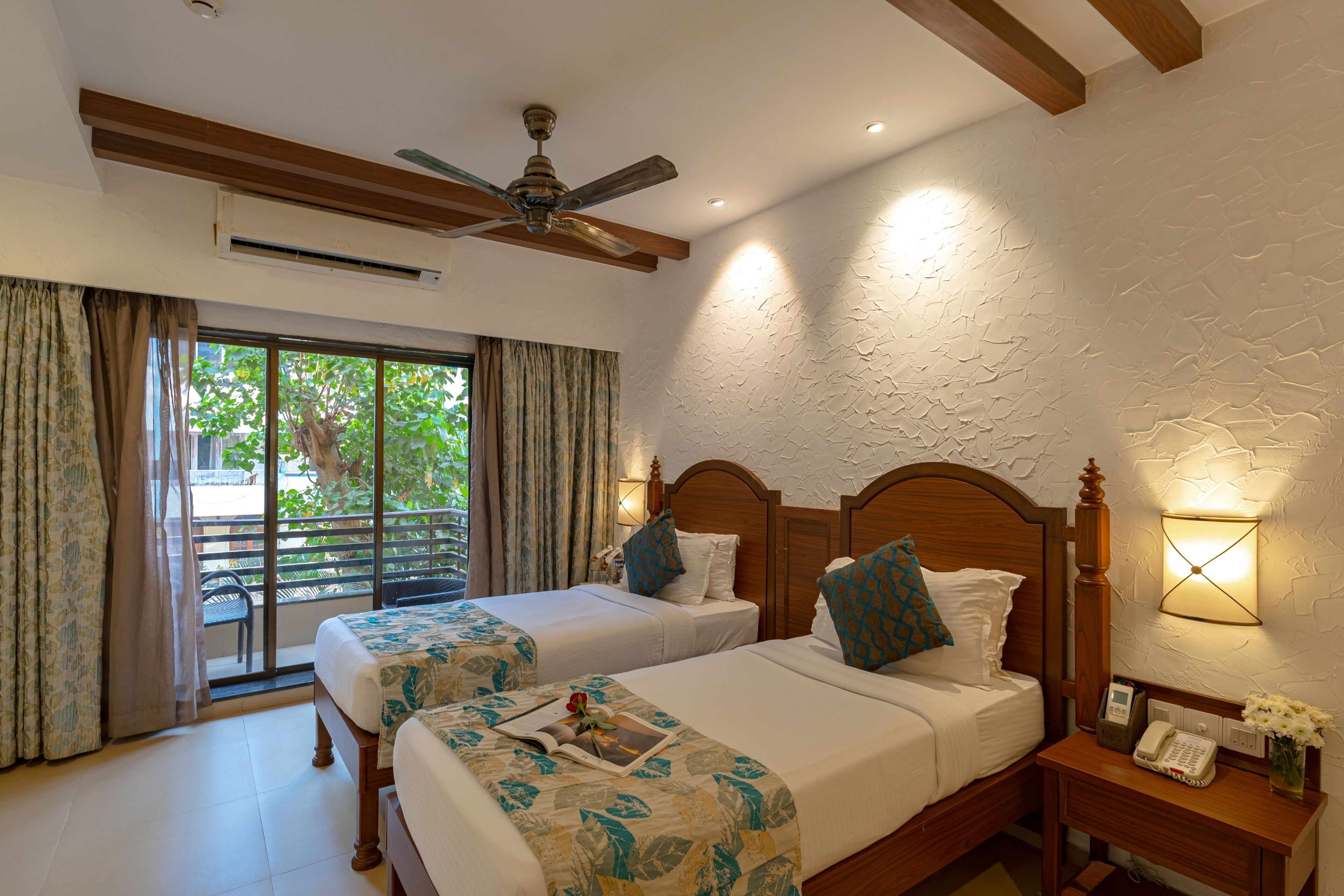 Une chambre d’hôtel avec deux lits, un ventilateur de plafond, un appareil de climatisation, une table de chevet avec un téléphone et un vase de fleurs, et une vue sur l’extérieur grâce à une porte-fenêtre coulissante.