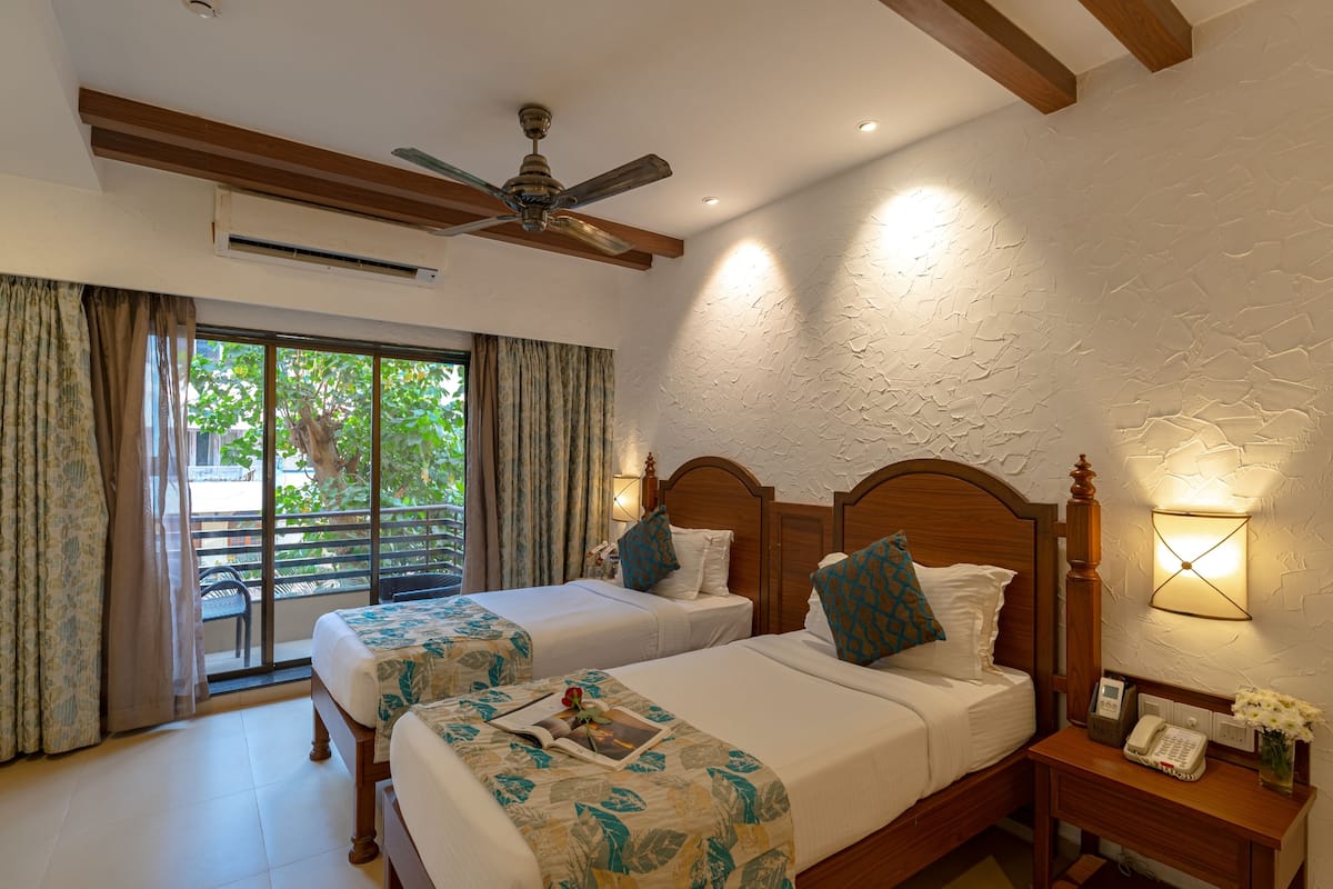 A hotel room with two beds, a ceiling fan, an air conditioning unit, a nightstand with a phone and a vase of flowers, and a view through a sliding glass door.