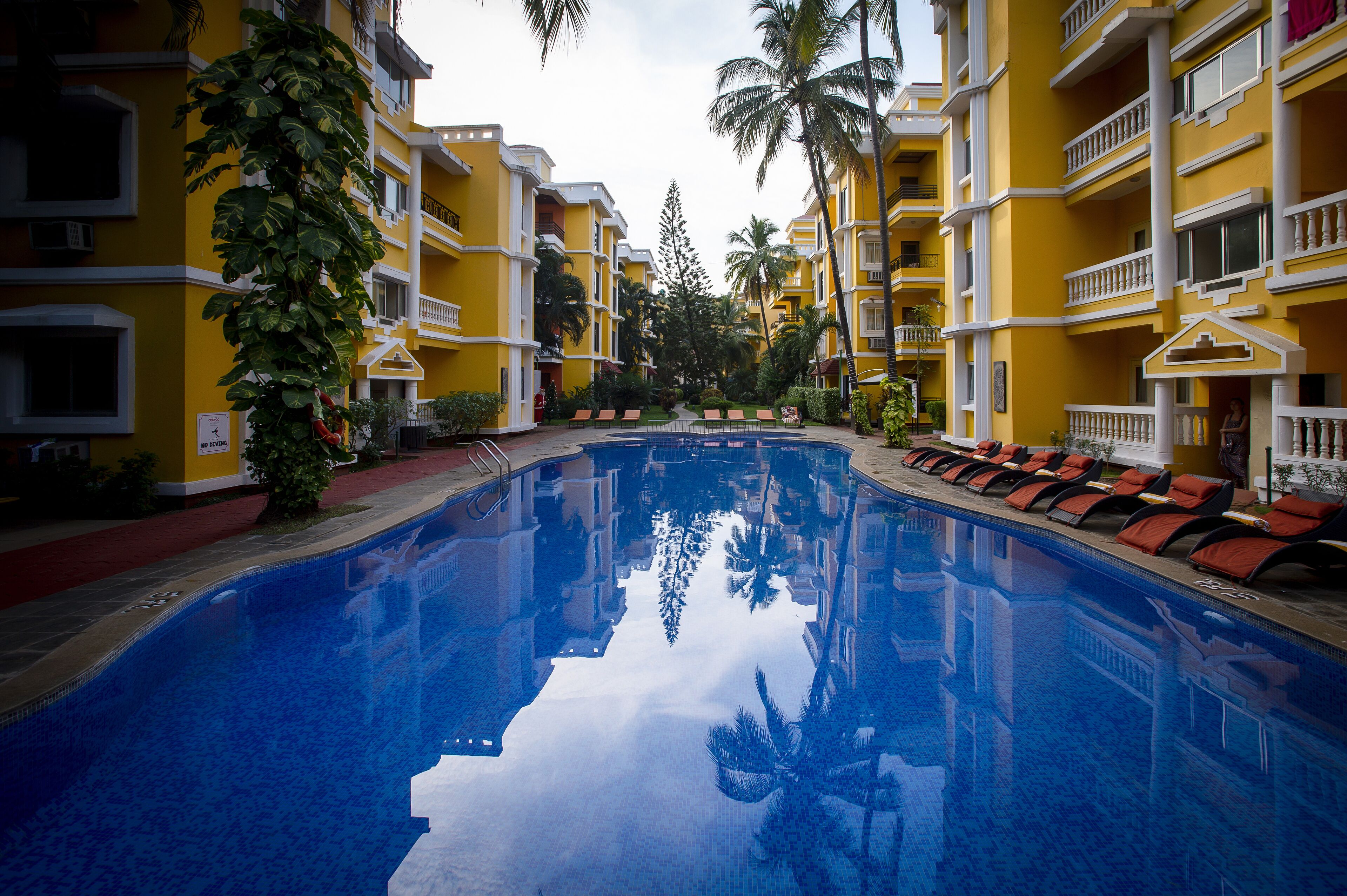 Una piscina con sillones de descanso y palmeras, rodeada de edificios amarillos con balcones.