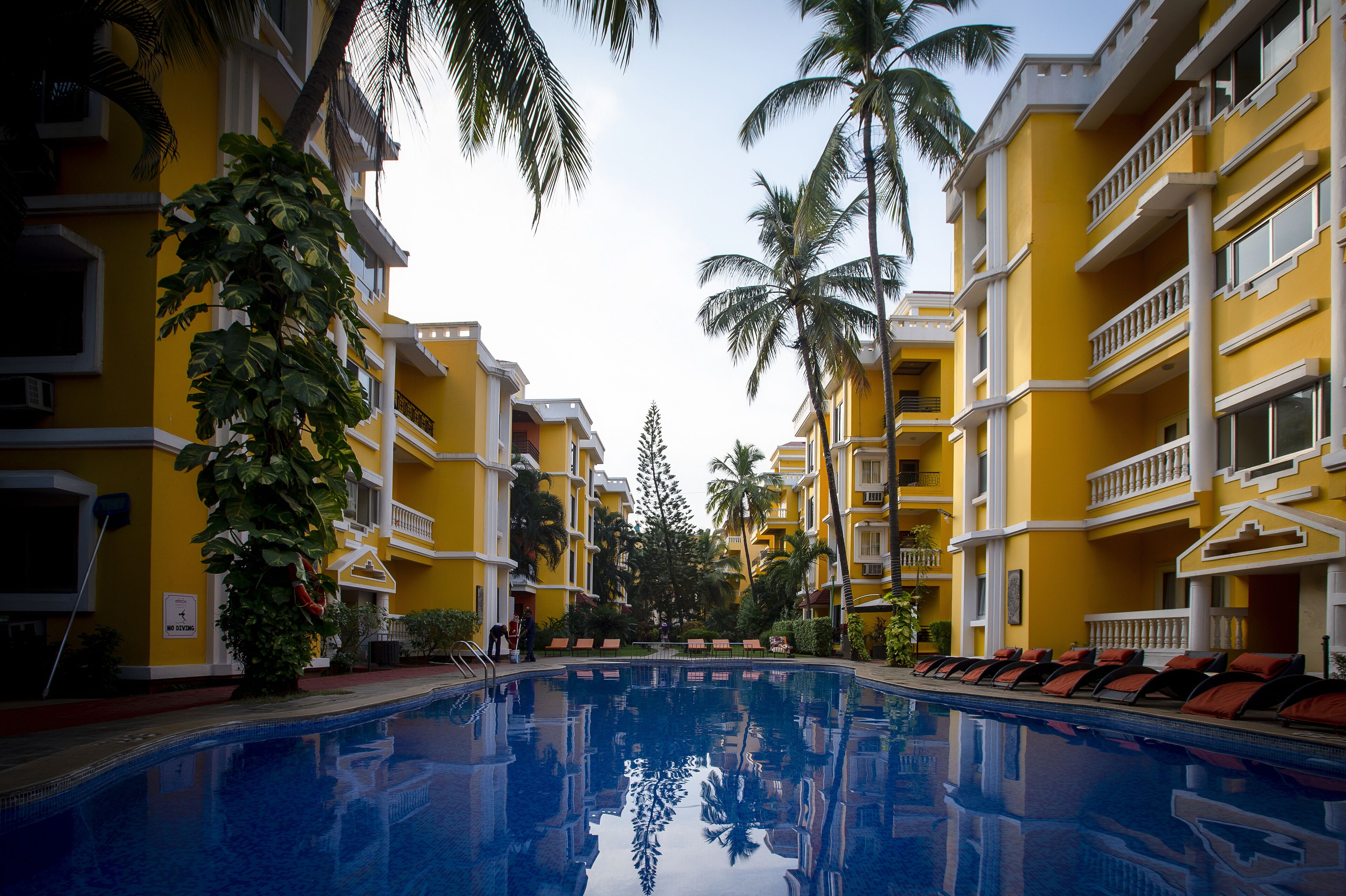 Un hôtel doté d’une piscine, entouré de bâtiments jaunes, de palmiers et de chaises longues rouges.