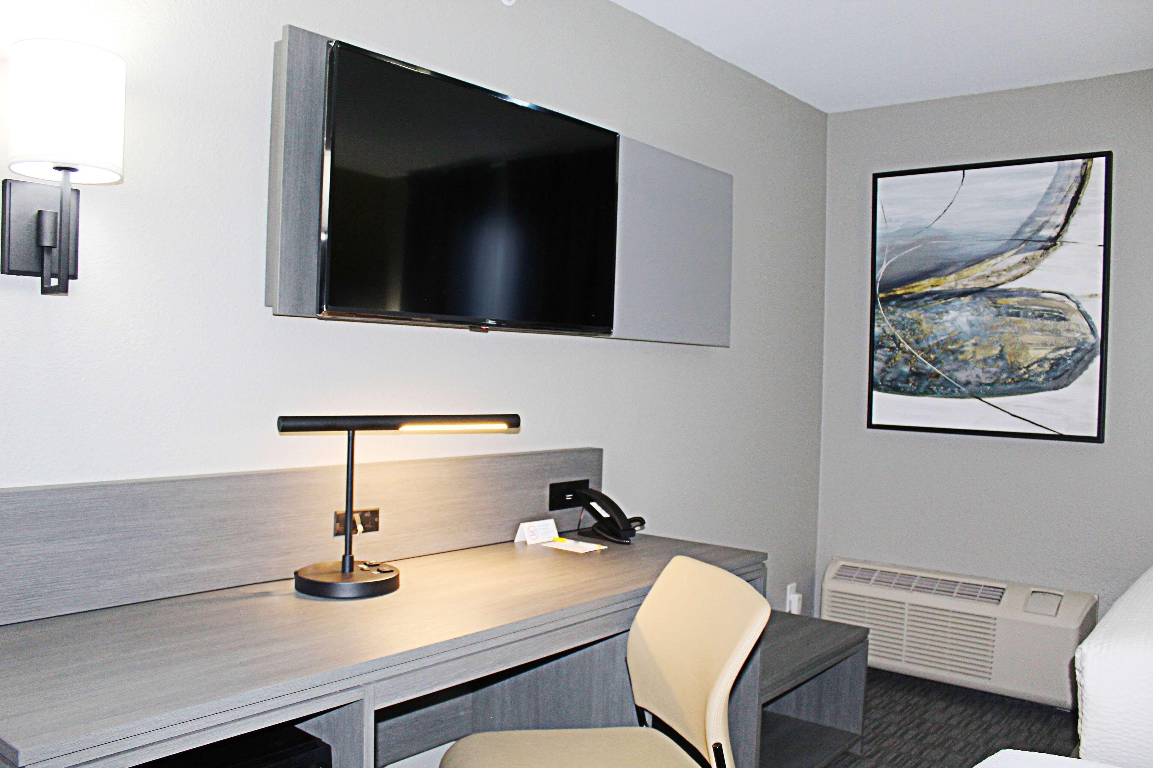 A hotel room with a desk, chair, flat-screen TV, artwork, and a wall-mounted lamp.