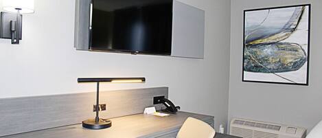 A hotel room with a desk, chair, flat-screen TV, artwork, and a wall-mounted lamp.
