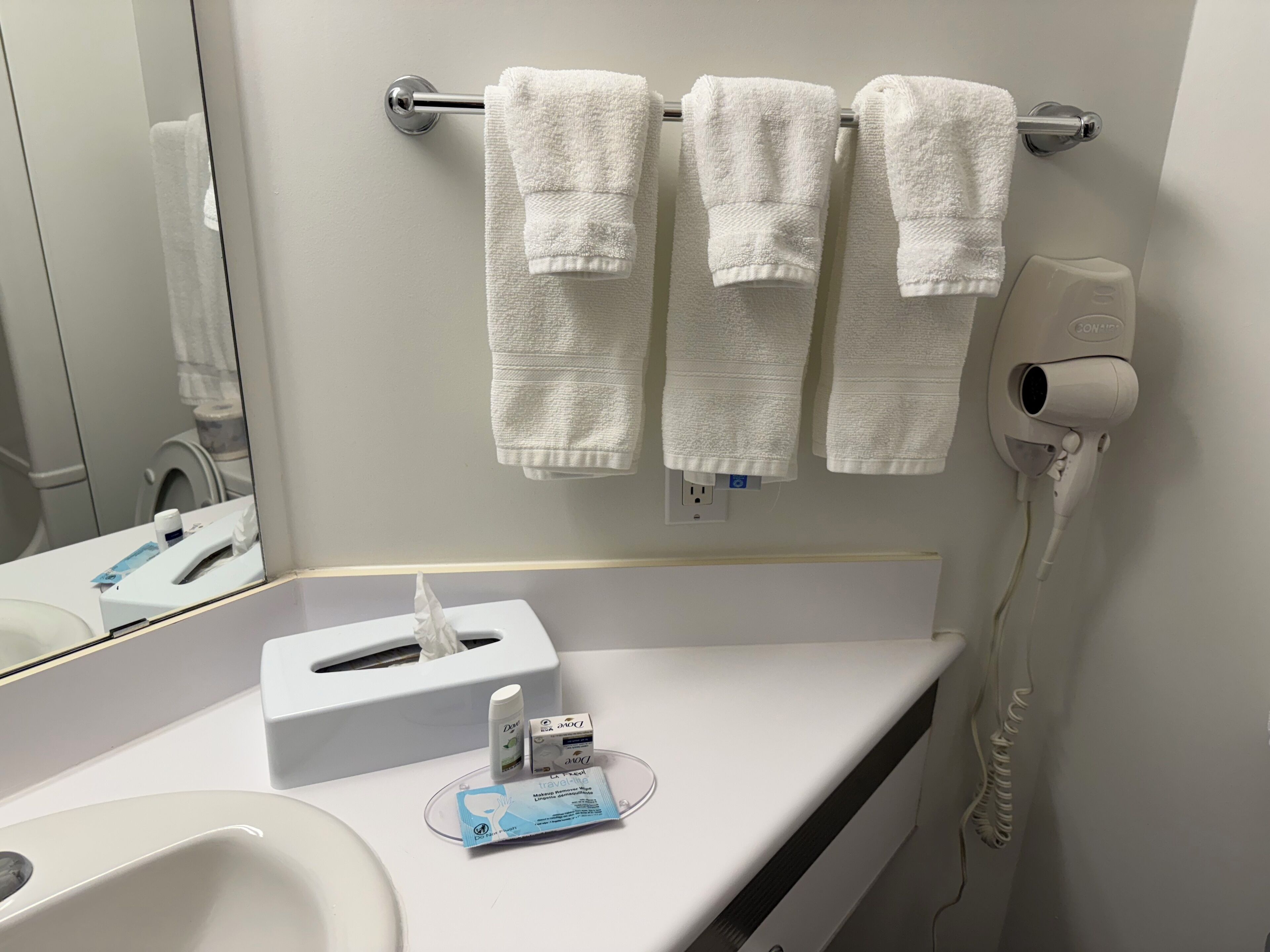 A bathroom with white towels hanging on a rack, a tissue box, a hand sanitizer, and a hair dryer plugged in.