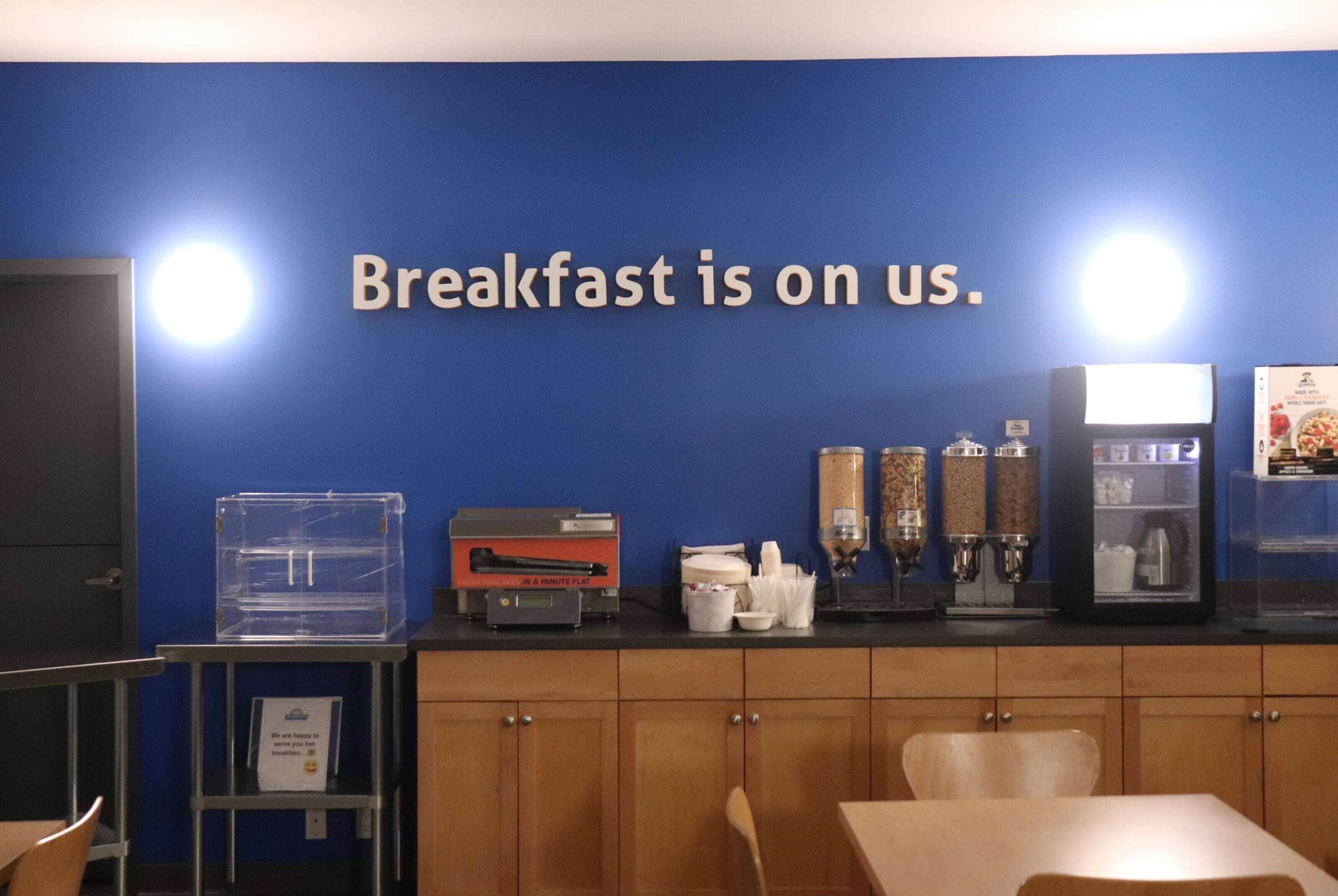 Een ontbijtruimte met een bord waarop staat 'Ontbijt van het huis'. Er is een koffiezetapparaat, een koelkast en een aanrecht met diverse spullen.