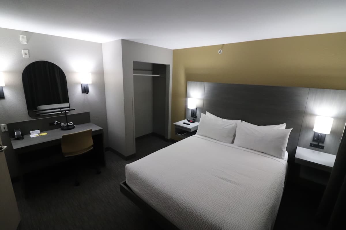 A hotel room with a bed, a desk, a chair, a wardrobe, and a nightstand with a lamp.