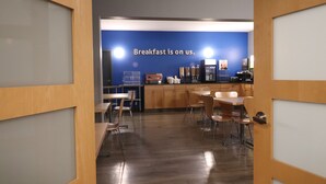 A modern breakfast area with a blue wall and a sign stating "Breakfast is on us." There are wooden tables and chairs, and a coffee machine is visible. - Days Inn by Wyndham Stony Plain (Stony Plain)