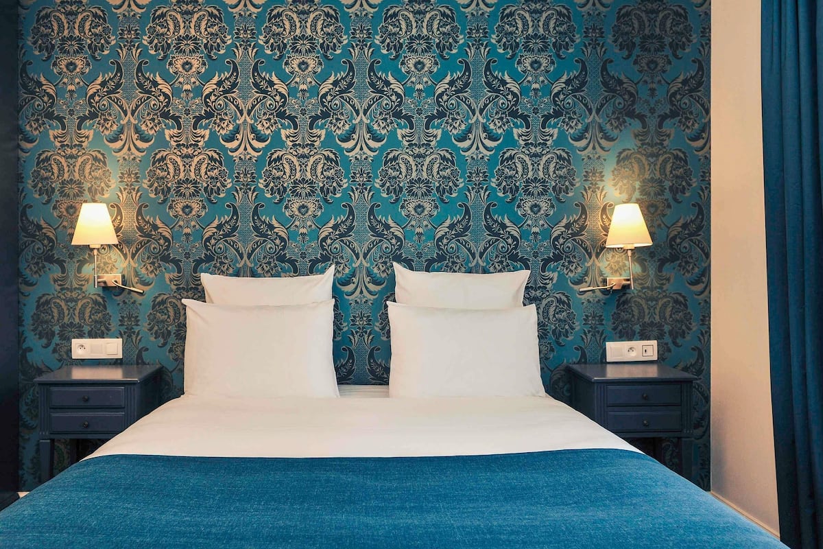 Un lit recouvert dâune couvre-lit bleue, deux oreillers blancs et un papier peint Ă motifs. Il y a deux tables de chevet, chacune Ă©quipĂ©e dâune lampe et dâune prise de courant.