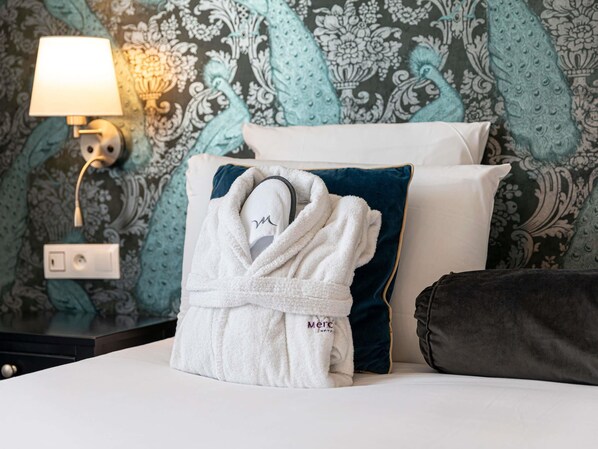 A neatly made bed with a white bathrobe, a dark blue pillow, and a white pillow. A wall-mounted lamp is visible on the left side of the bed. - Mercure Paris Saint Cloud Hippodrome (Saint-Cloud)