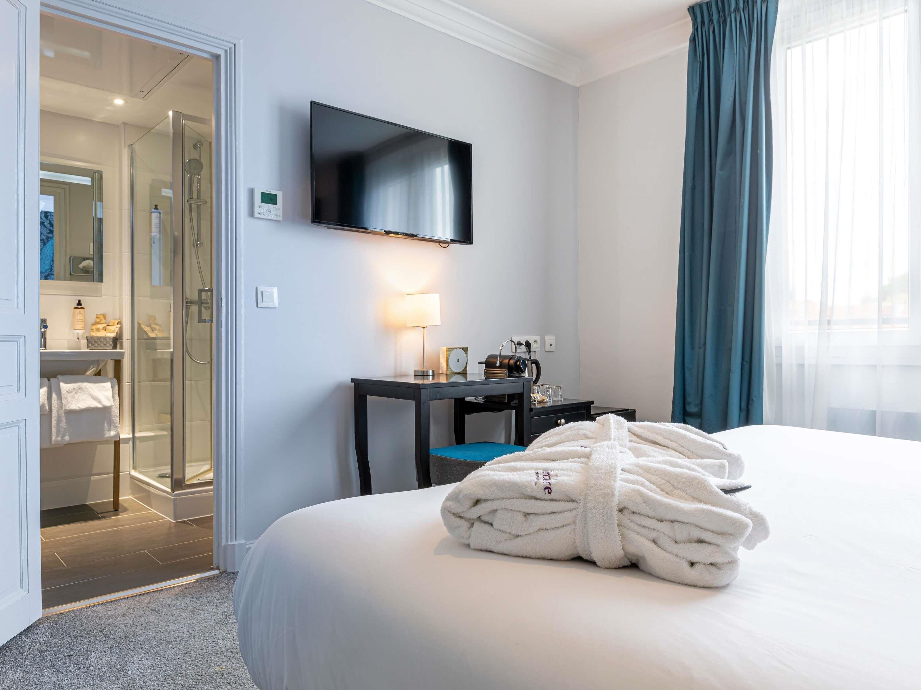 A hotel room with a bed, a TV mounted on the wall, a desk with a lamp, and a bathroom visible through an open door.