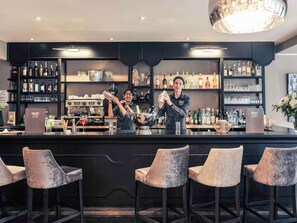 A bar area with a bartender preparing drinks, a coffee machine, and a variety of bottles and glasses on shelves.