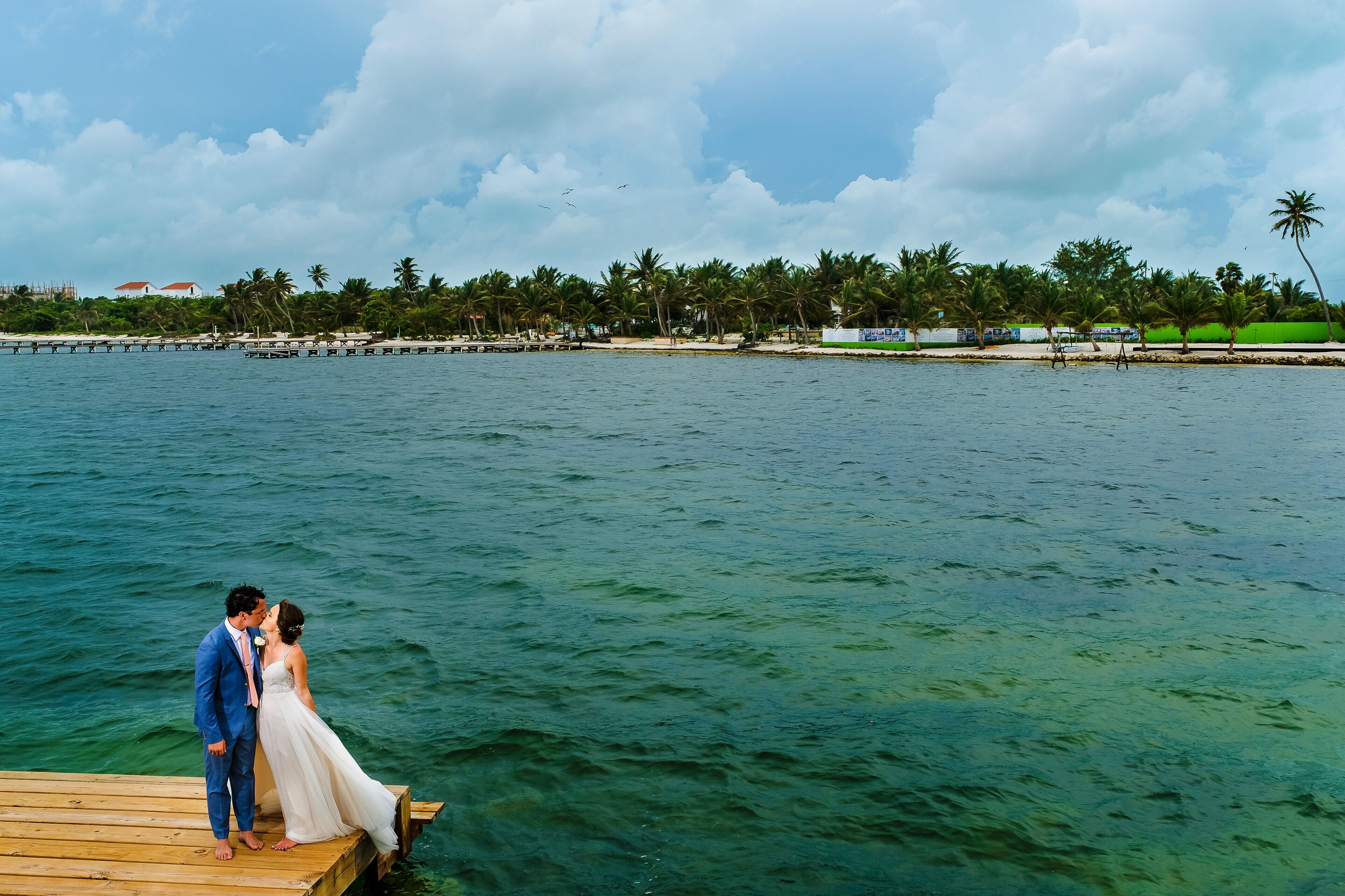 Una pareja en un muelle de madera al lado del agua, con una playa y palmeras al fondo.
