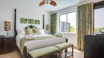 Quarto de hotel com cama, um banquinho, ventilador de teto, janela com cortinas e obras de arte na parede.