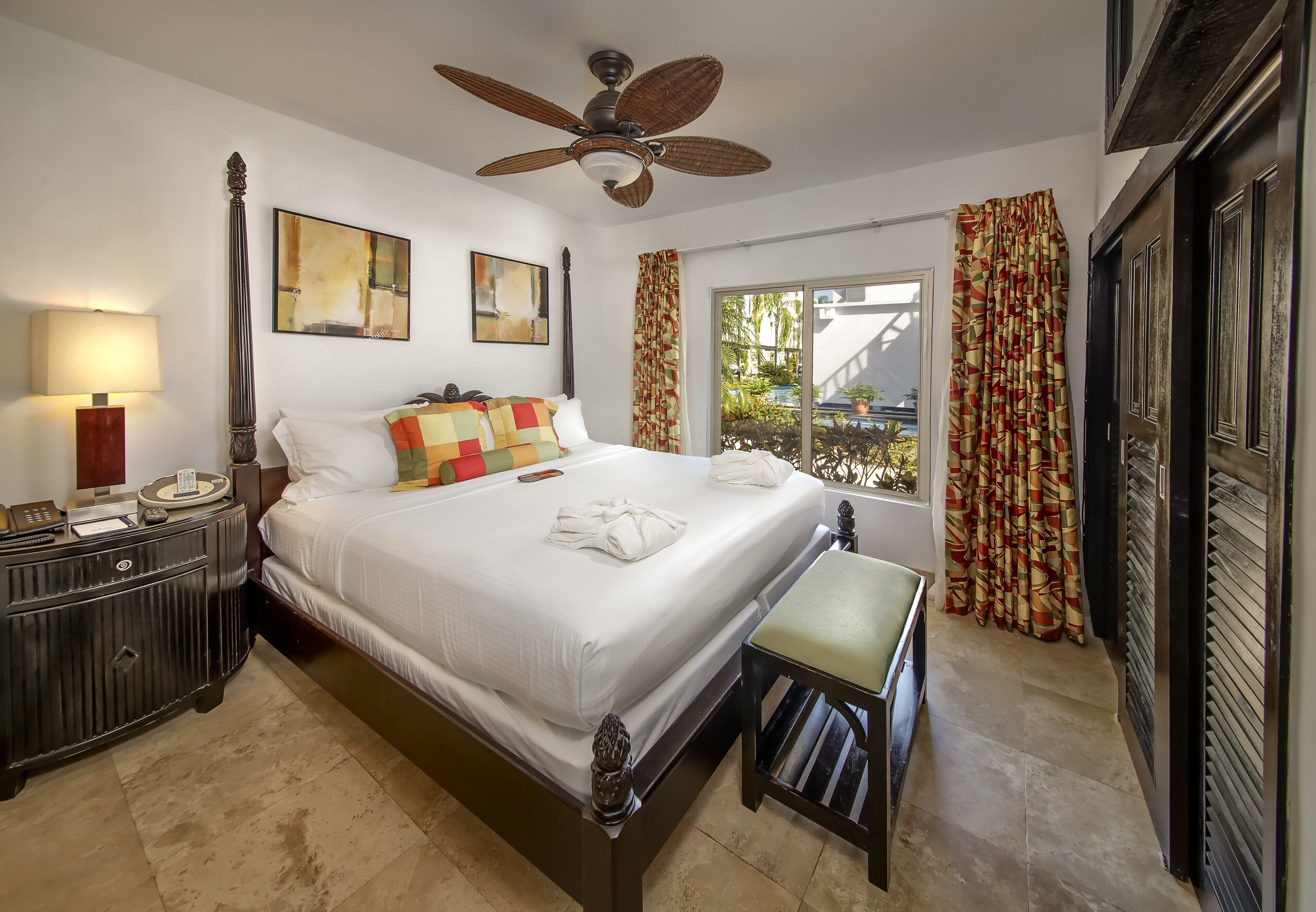 A hotel room with a large bed, a bedside table with a lamp, a ceiling fan, and a window with curtains.