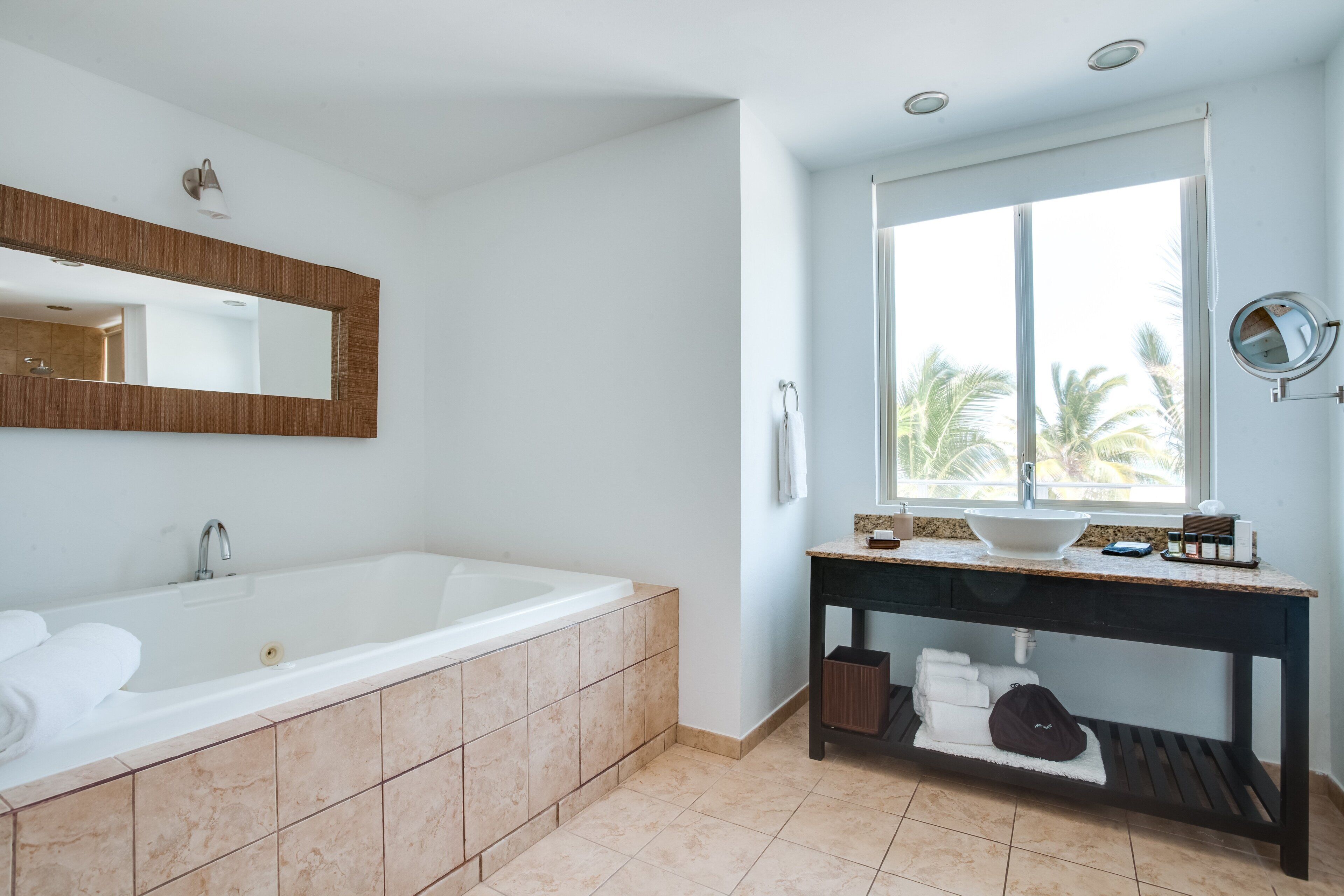 A modern bathroom with a large bathtub, a vanity with a sink, and a window providing a view of palm trees.