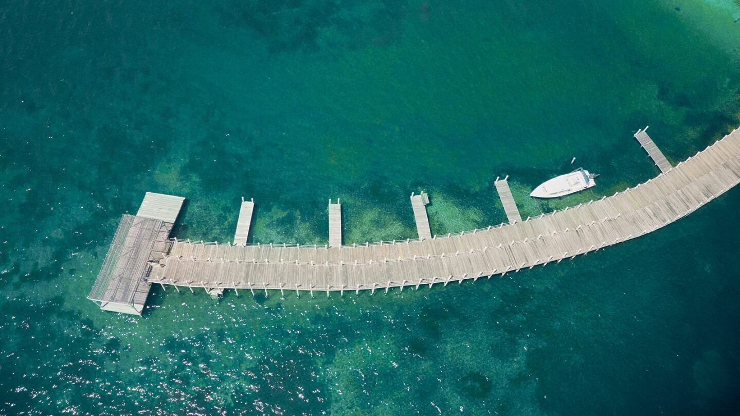 An aerial view of a long, straight pier extending into clear turquoise water, with a boat docked at the end and a small dock on the left.