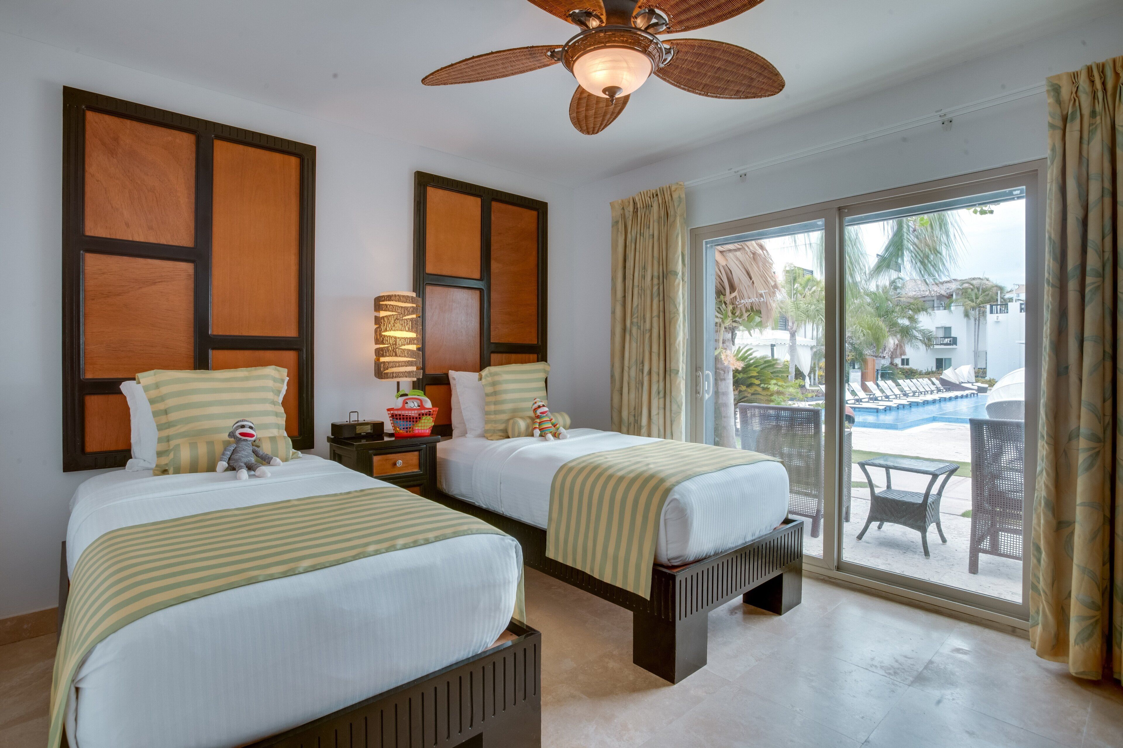 A hotel room with two beds, a ceiling fan, a large window with a view of a pool and palm trees, and a small table with chairs outside.