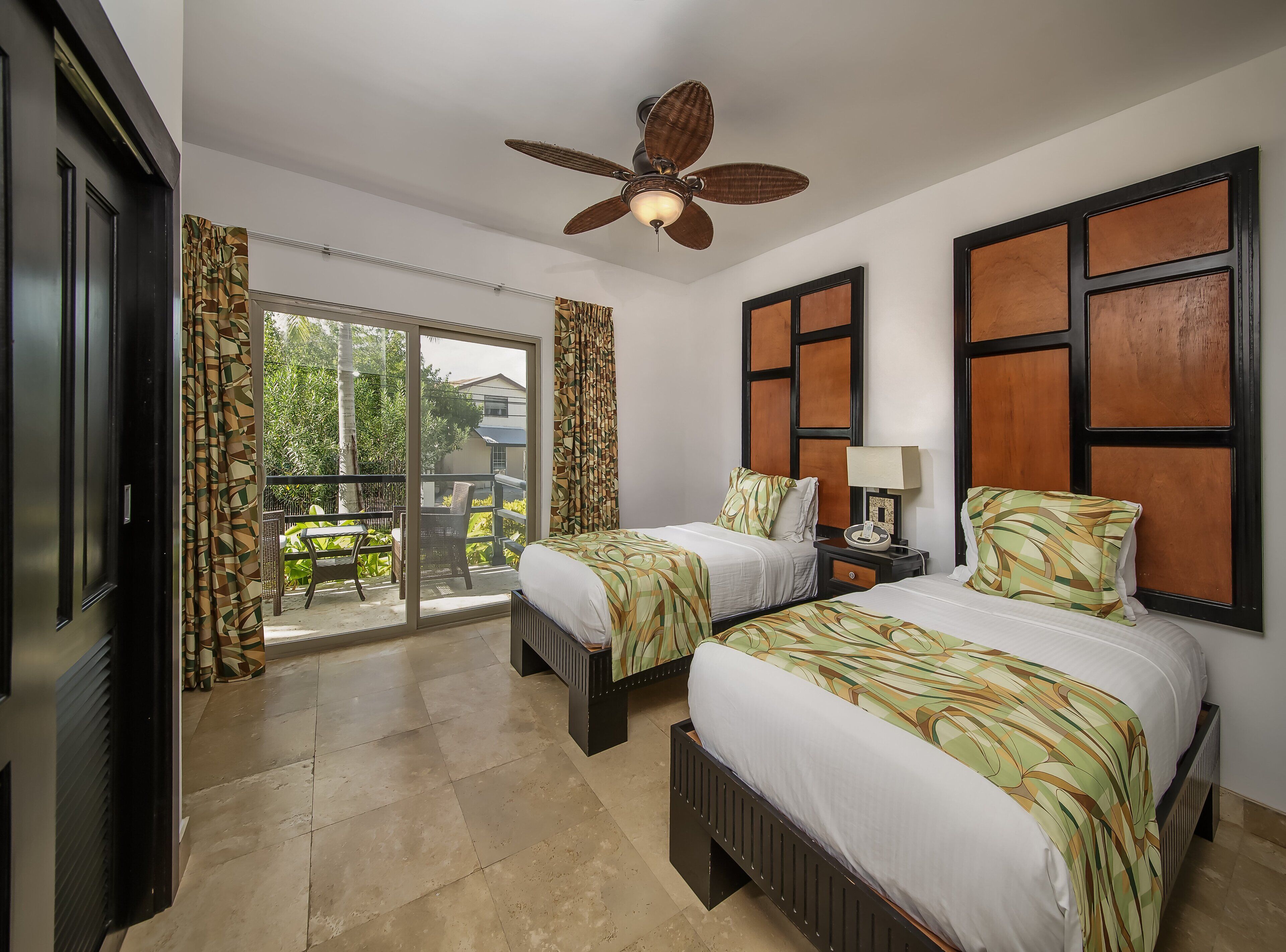 A hotel room with two beds, a ceiling fan, a sliding glass door to the balcony, and a nightstand with a lamp.