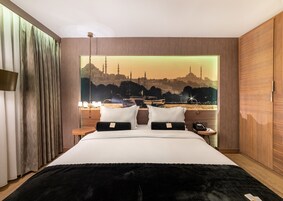 A hotel room with a large bed, a nightstand, a lamp, and a view of a cityscape with buildings and a mosque.