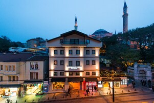 A multi-story building with a balcony, a mosque with a minaret in the background, and a street with shops and pedestrians.