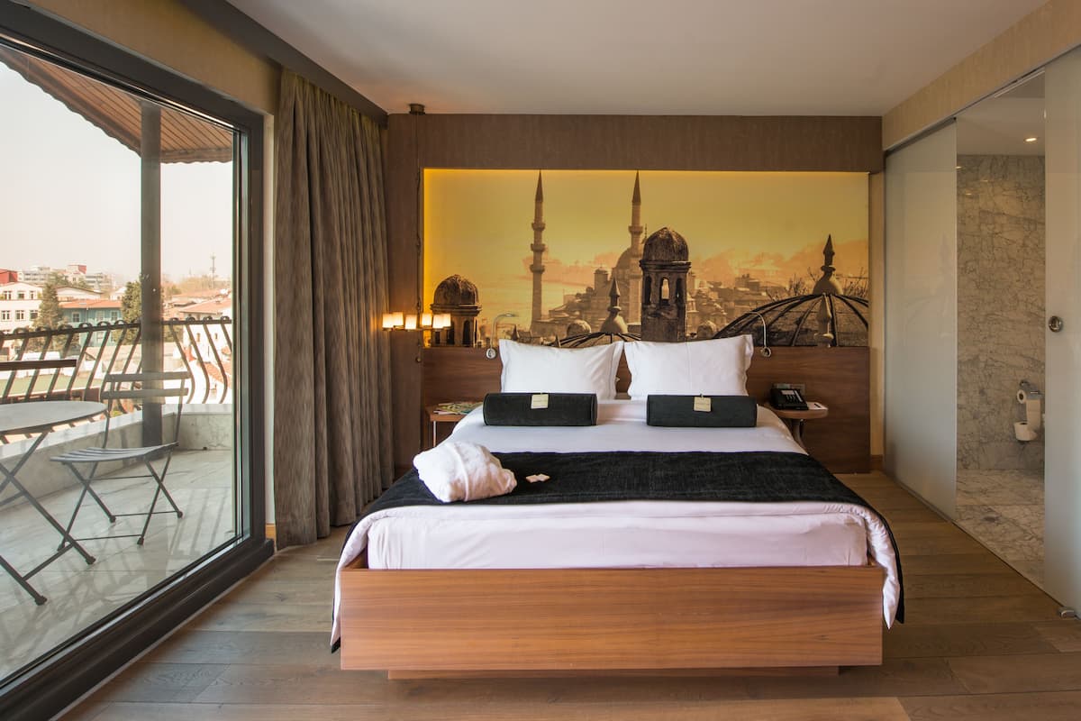 Une chambre d’hôtel avec un grand lit, un balcon offrant une vue, une fresque représentant un paysage urbain sur le mur et un plancher en bois.