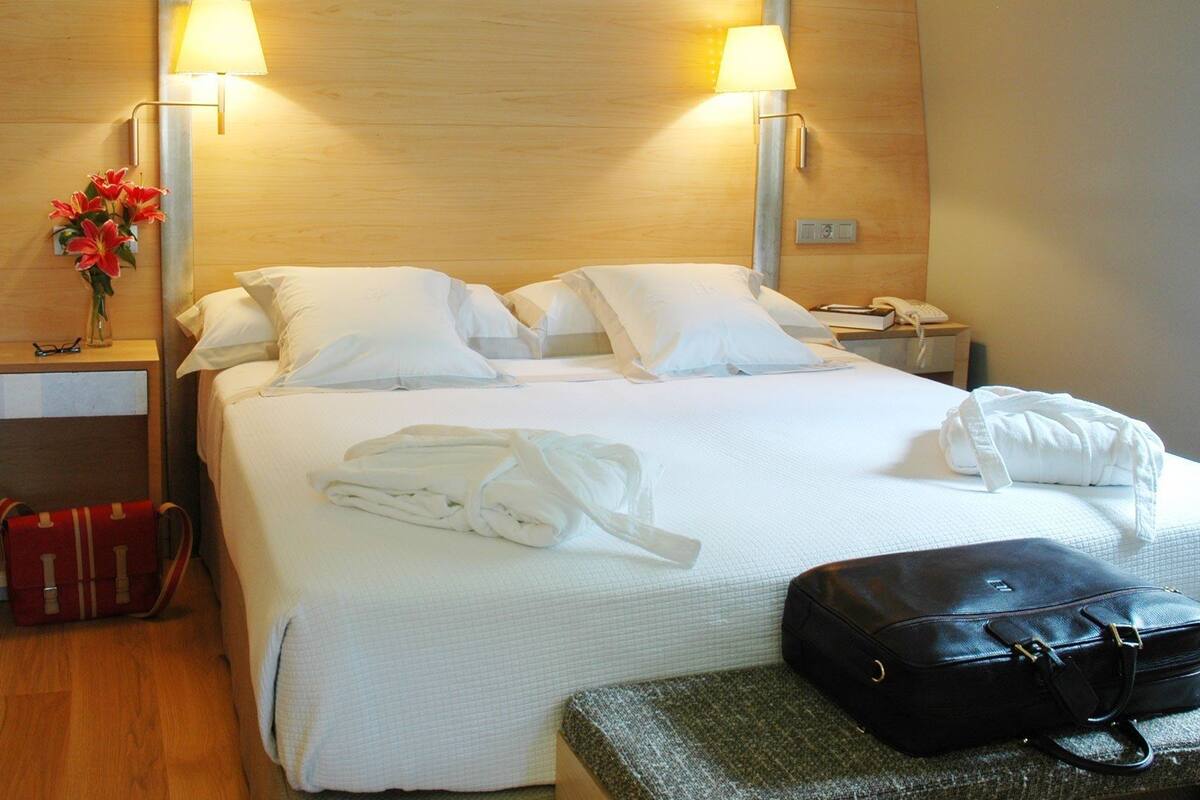 Une chambre d’hôtel avec un lit bien fait, deux lampes de chevet, un téléphone sur la table de chevet, une valise rouge, un sac à main noir et un vase avec des fleurs sur la table de chevet.