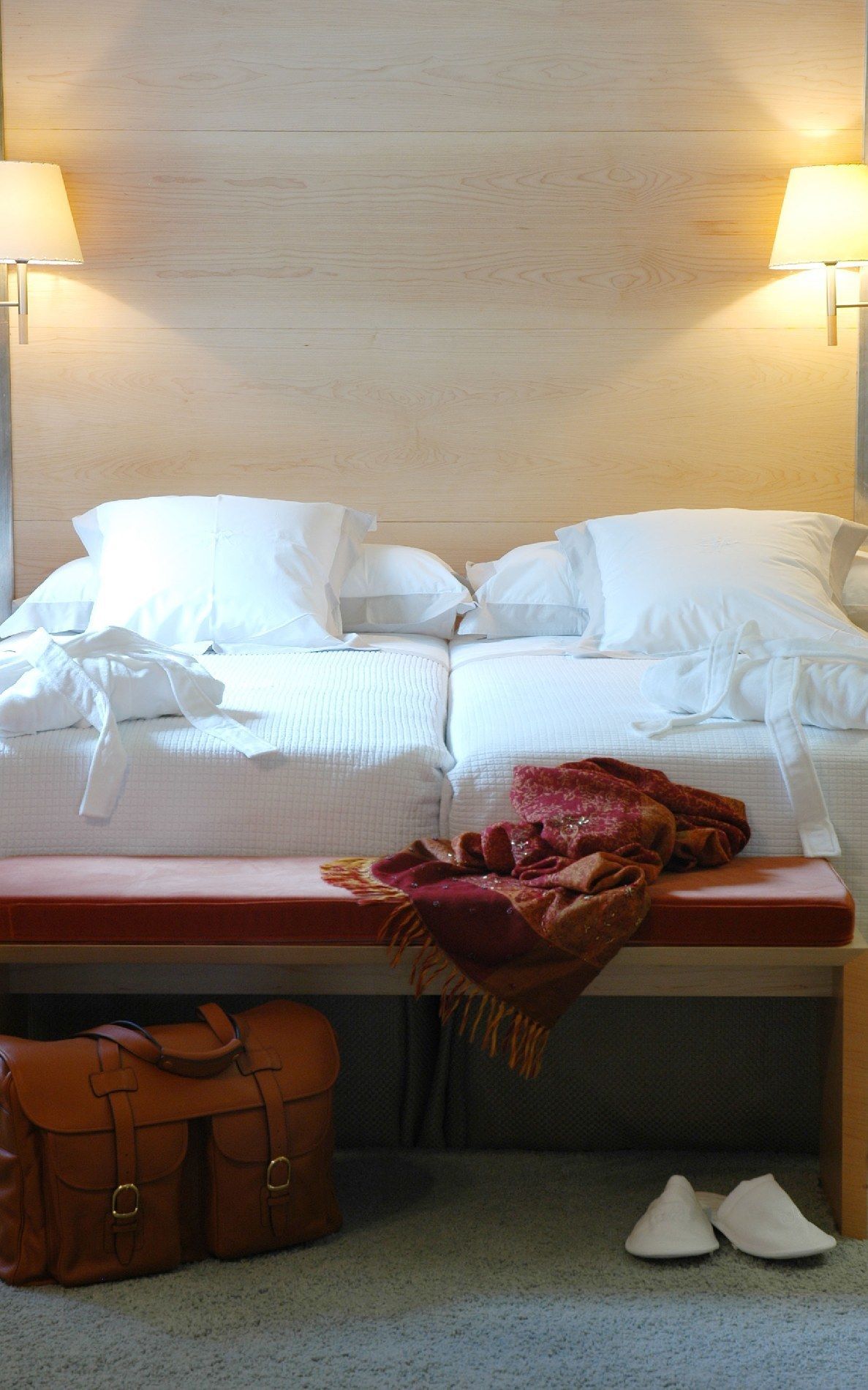 A neatly made bed with white linens and pillows, a wooden headboard, and a bedside table with a red blanket and brown leather bags.