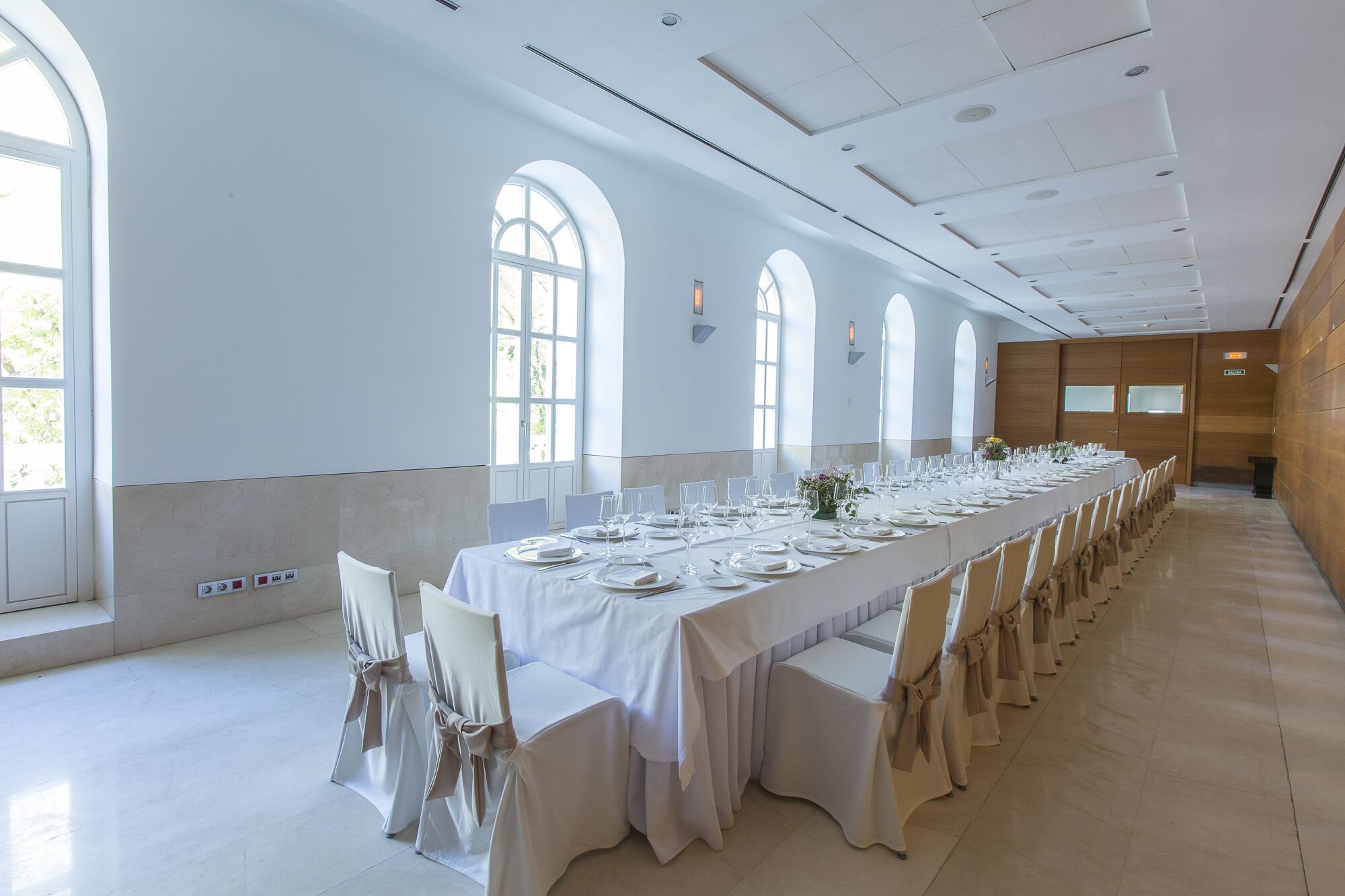 Una larga mesa de banquete dispuesta con manteles blancos y sillas, amplias ventanas en arco y una pared de madera.