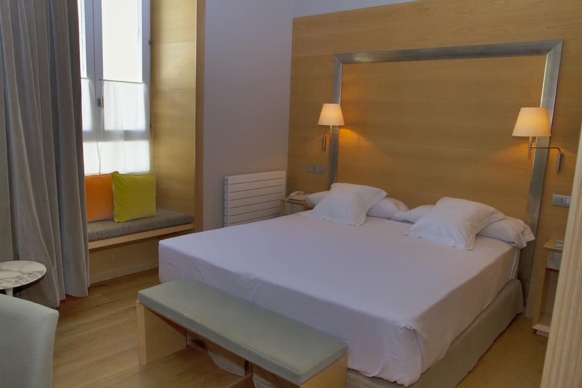 Une chambre d’hôtel avec un grand lit, deux lampes de chevet, un banc, une petite table et une fenêtre avec des rideaux.
