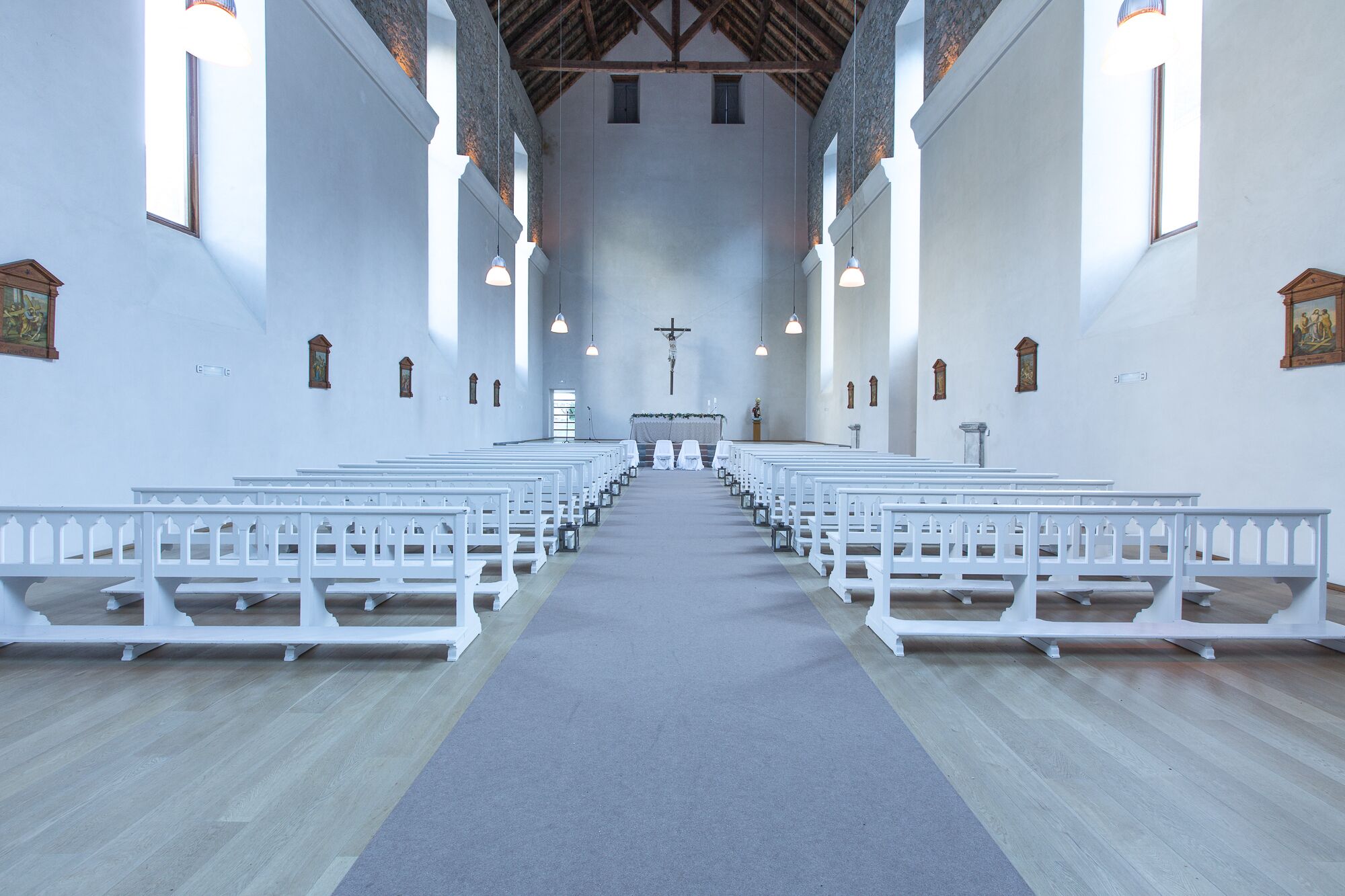 Interior de una iglesia con bancas blancas dispuestas en filas, un pasillo central que conduce al altar, y una cruz en lo alto.