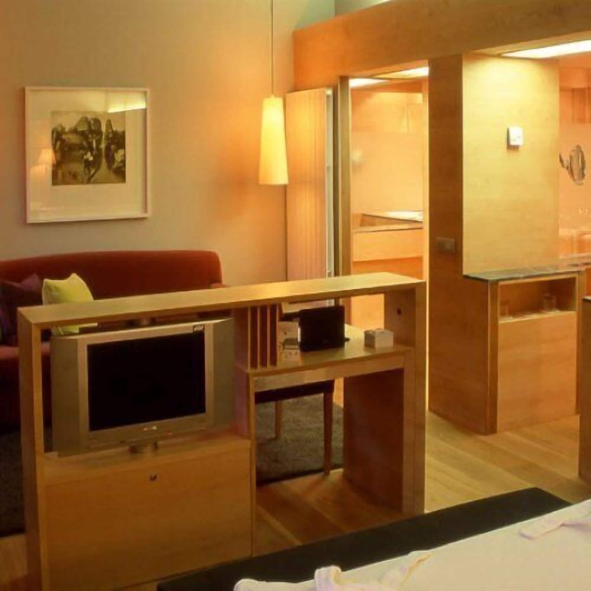 Kompakti hotellihuone, jossa on puinen työpöytä, televisio, punainen sohva ja seinällä oleva kehystetty kuva.