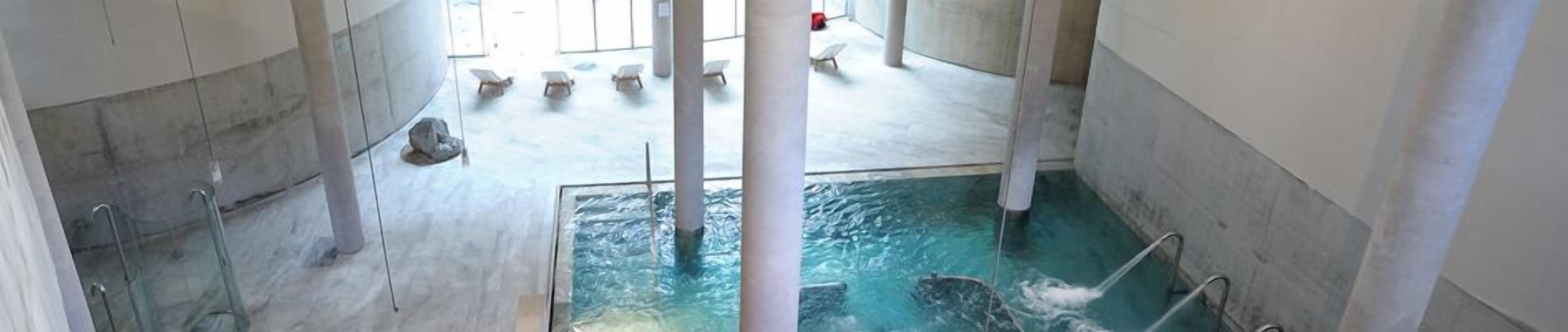 Een overdekt zwembad met helder water, meerdere banen en ligbedden op de achtergrond.