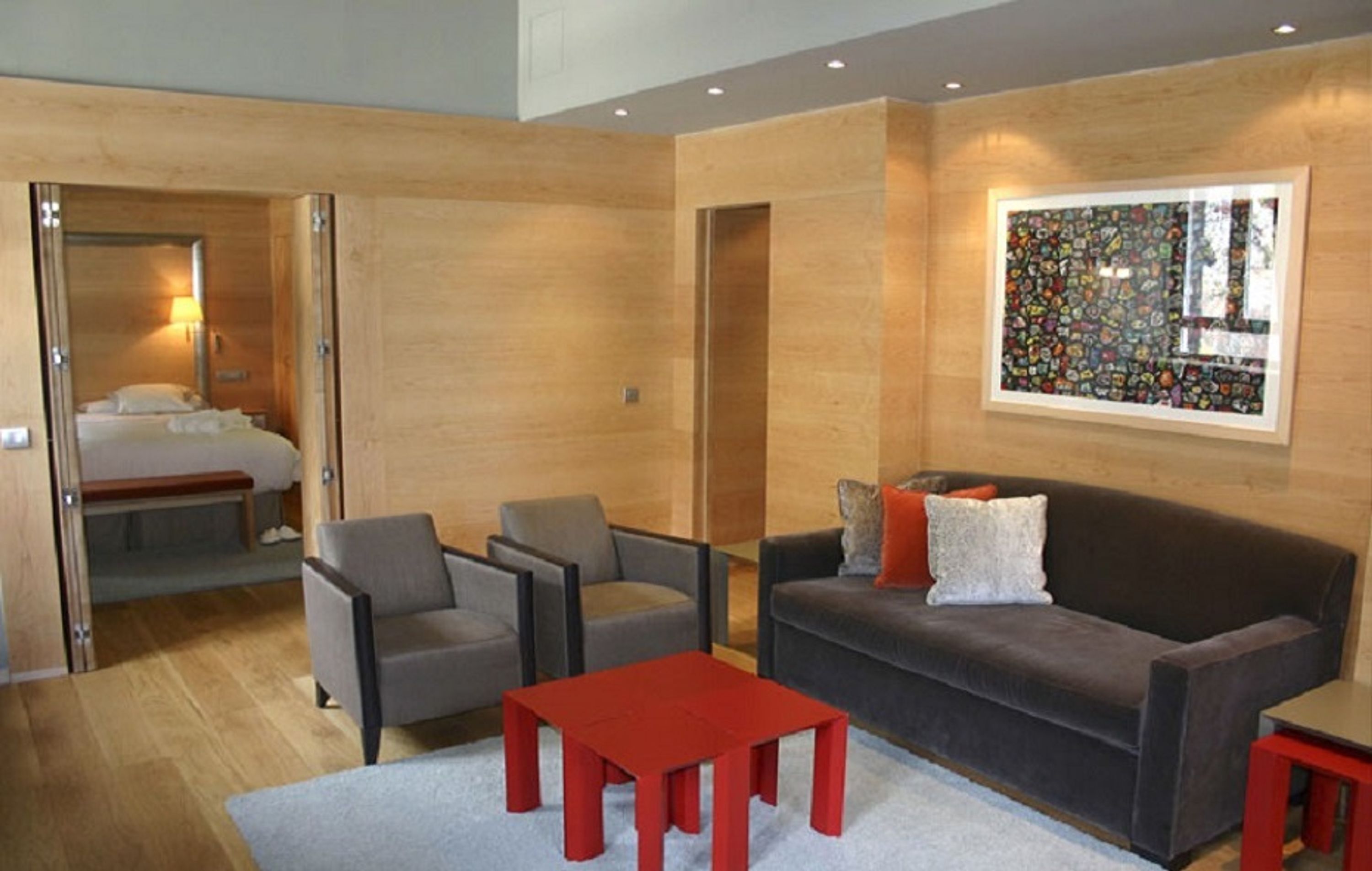 Quarto de hotel moderno com decoração em madeira, uma janela grande com uma obra de arte colorida, um sofá, duas poltronas, uma mesinha vermelha e uma cama com um banco.