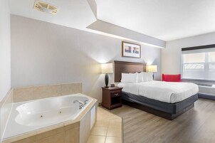 Habitación de hotel con una bañera grande, una cama con ropa blanca, una mesita de noche con lámparas y una ventana con persianas.