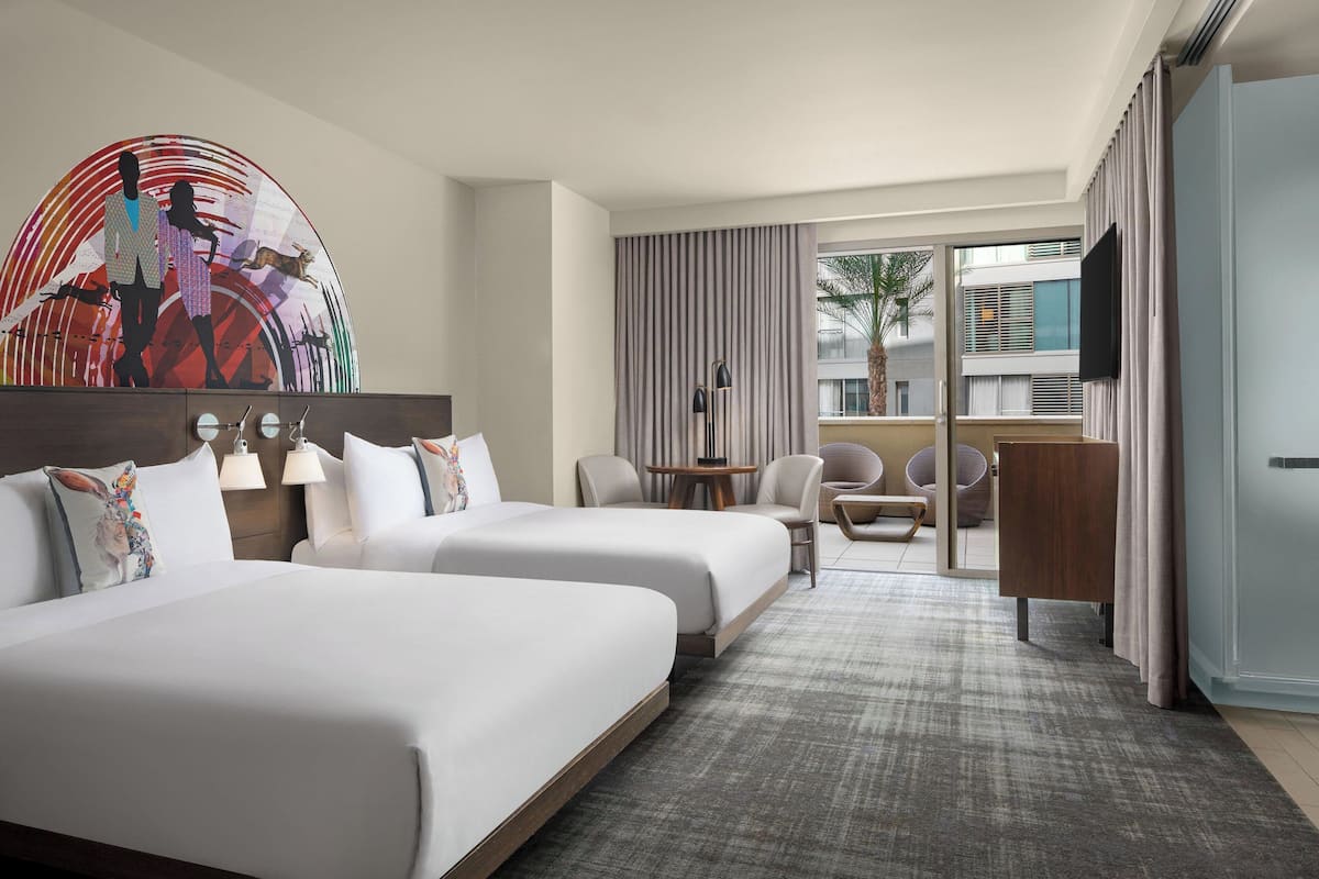 Habitación de hotel con dos camas, un escritorio, sillas, televisor y un amplio ventanal con vistas a edificios y palmeras.