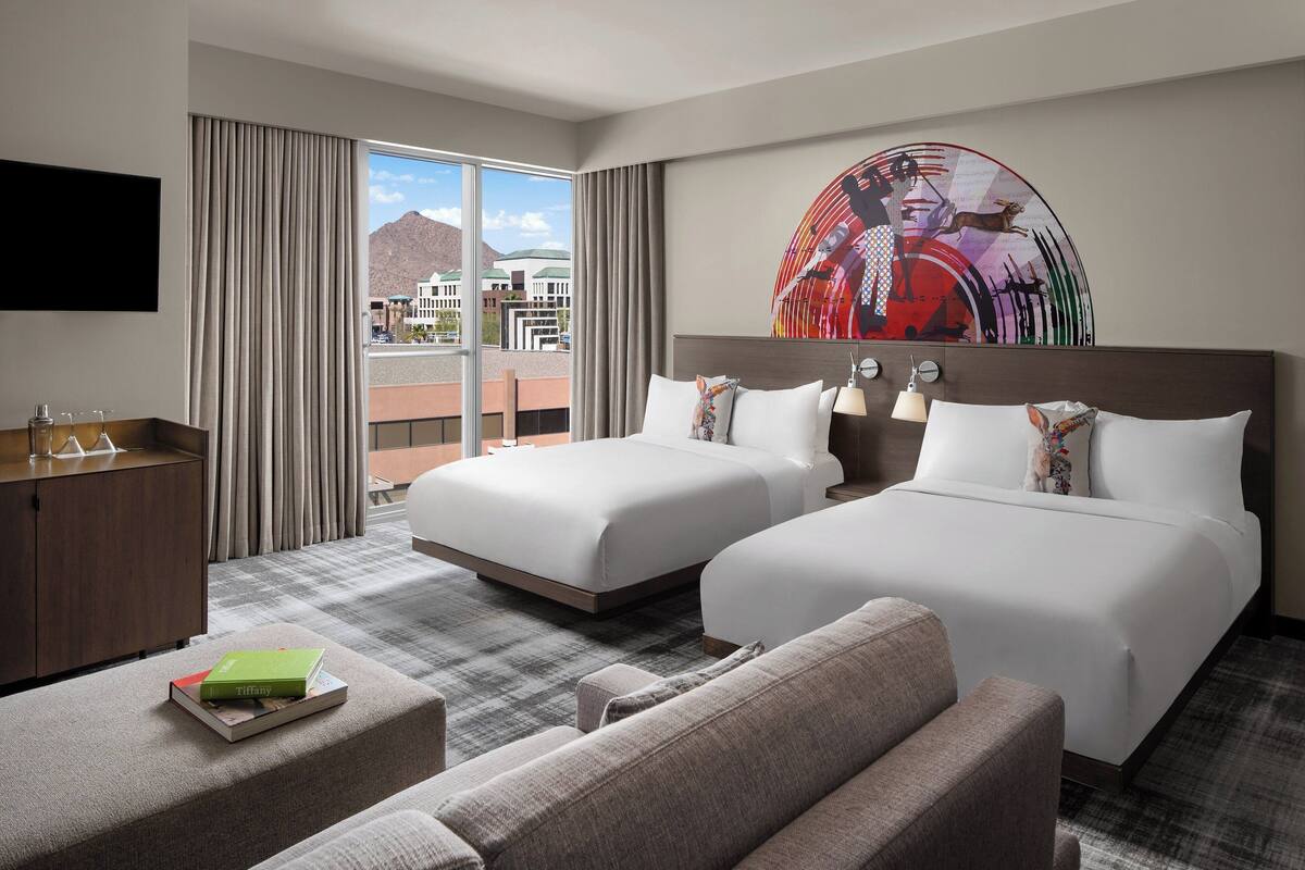 Habitación de hotel con dos camas, un sofá, un escritorio y un ventanal con vistas a edificios y montañas.