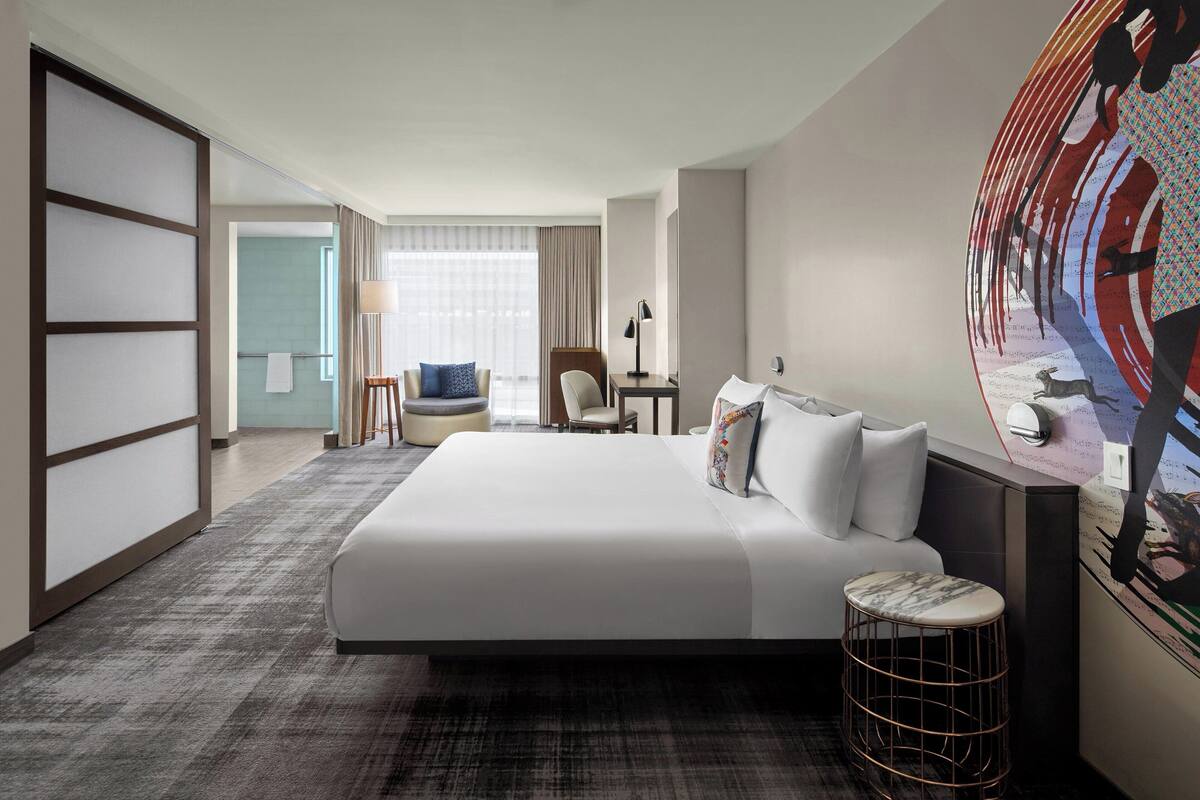 Una habitación de hotel con una cama grande, una zona de estar con un sillón y reposapiés, un escritorio con lámpara y un mural grande en la pared.