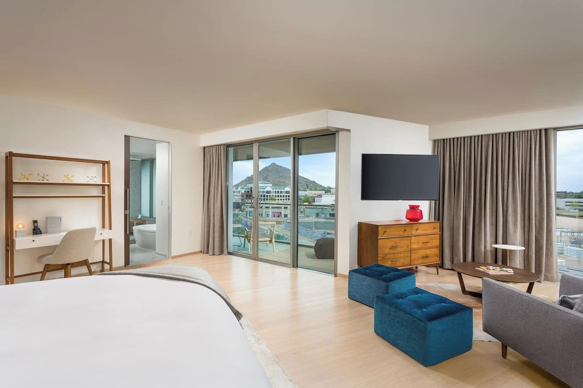 Una habitación de hotel moderna con una cama grande, un escritorio, un sofá azul, un televisor y un balcón con vista a la ciudad.