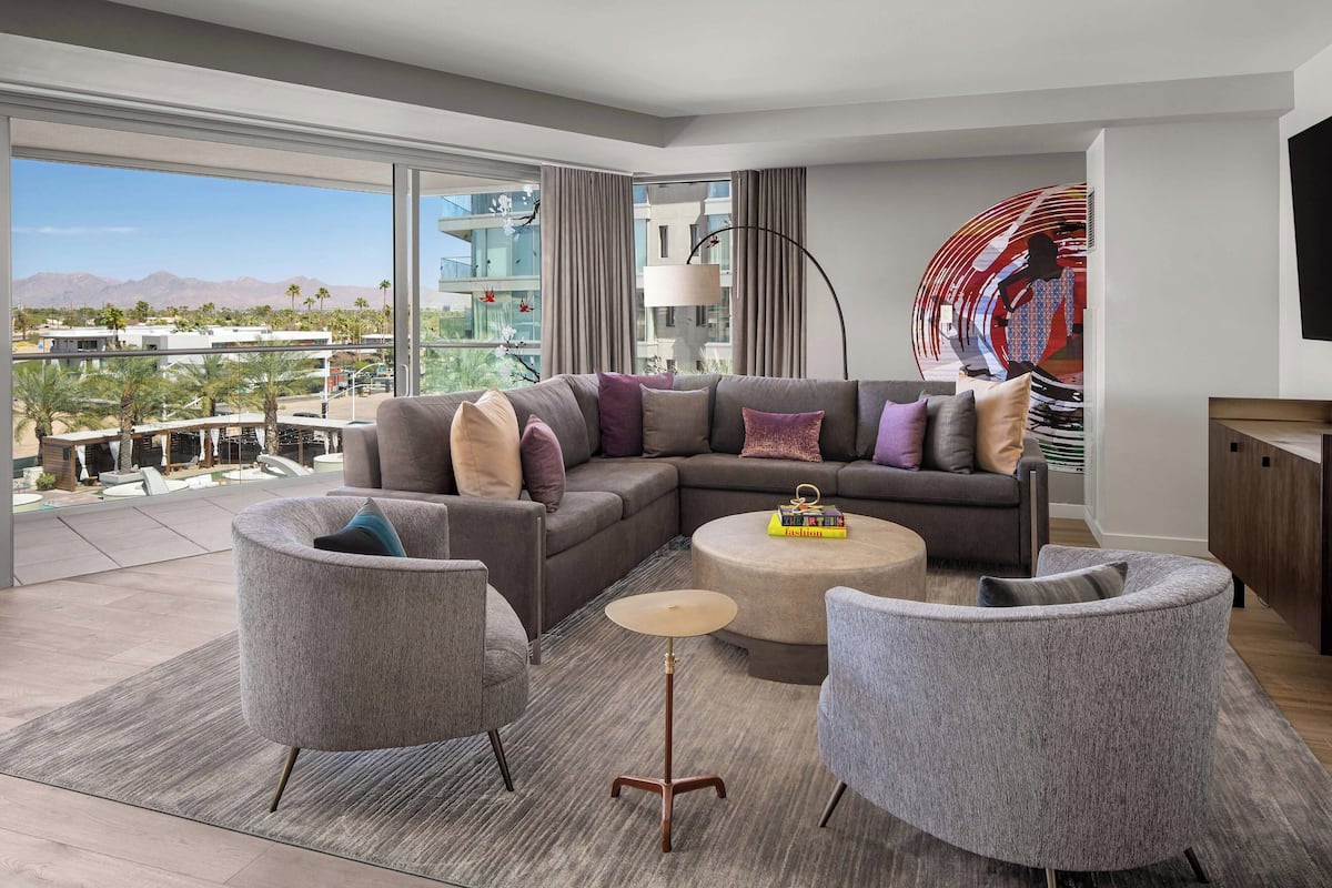 Una sala de estar moderna con un sofá seccional, dos sillones, una mesa de centro y un ventanal con vistas a palmeras y edificios.
