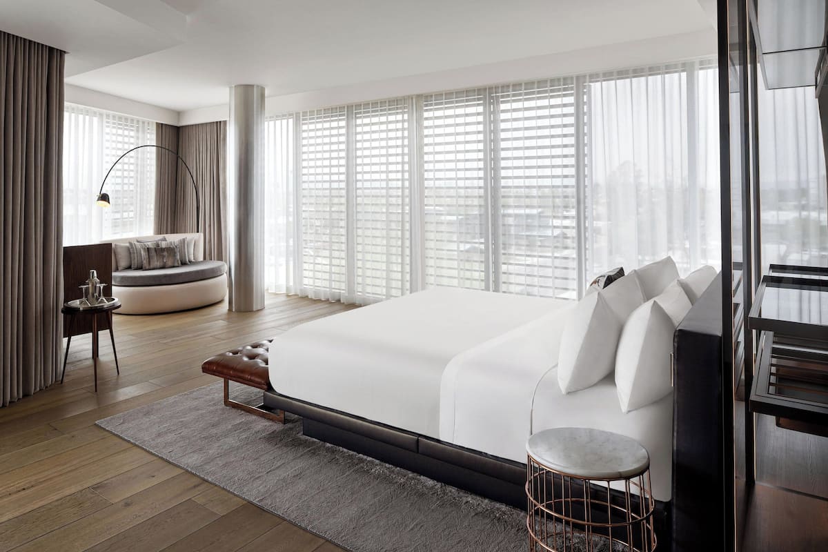 Un dormitorio amplio con una cama grande, una zona de estar con sofá y sillón, y vistas de la ciudad a través de amplias ventanas.