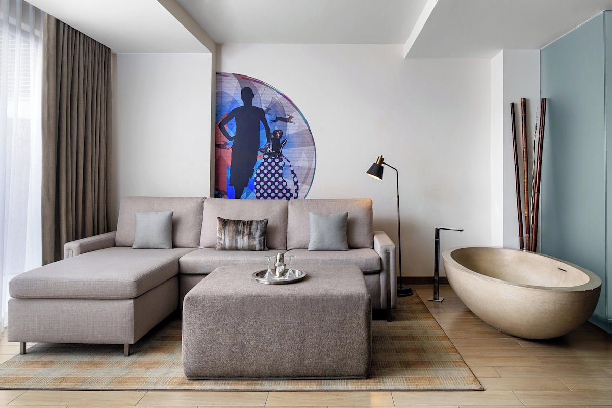 Una sala de estar moderna con un sofá modular, un otomán grande, una obra de arte en la pared, una lámpara de pie y una bañera amplia.