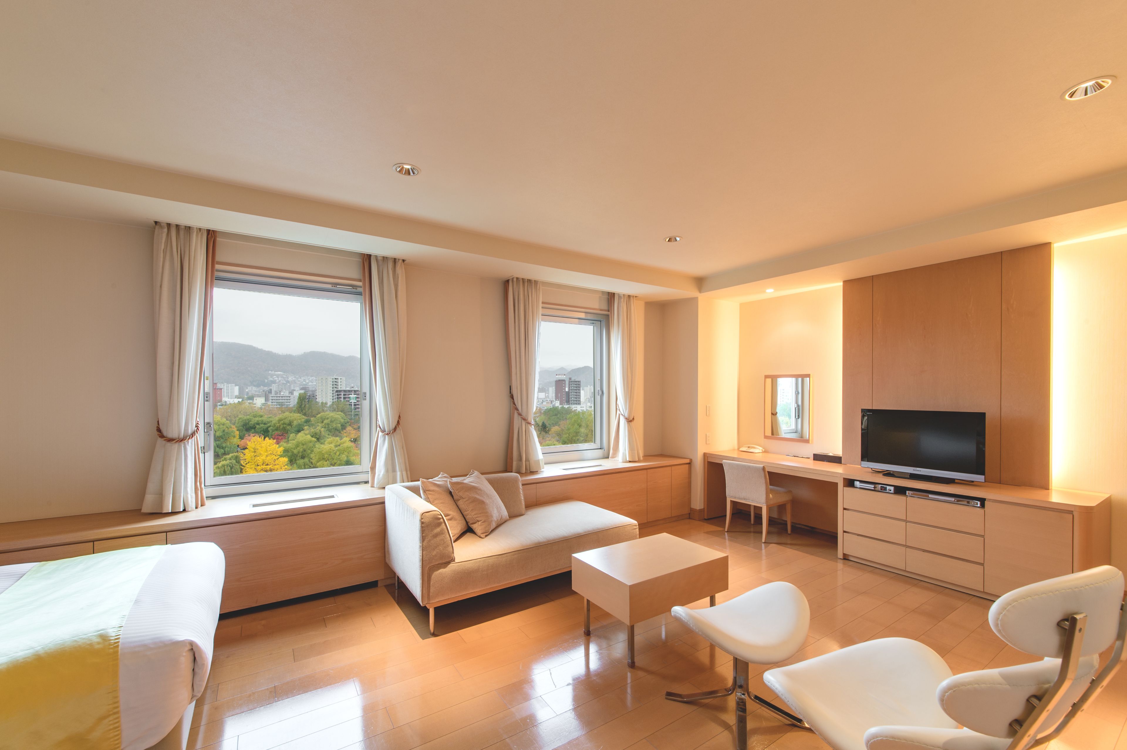 A modern hotel room with a sofa, a flat-screen TV, a desk, and a view of the city through large windows.