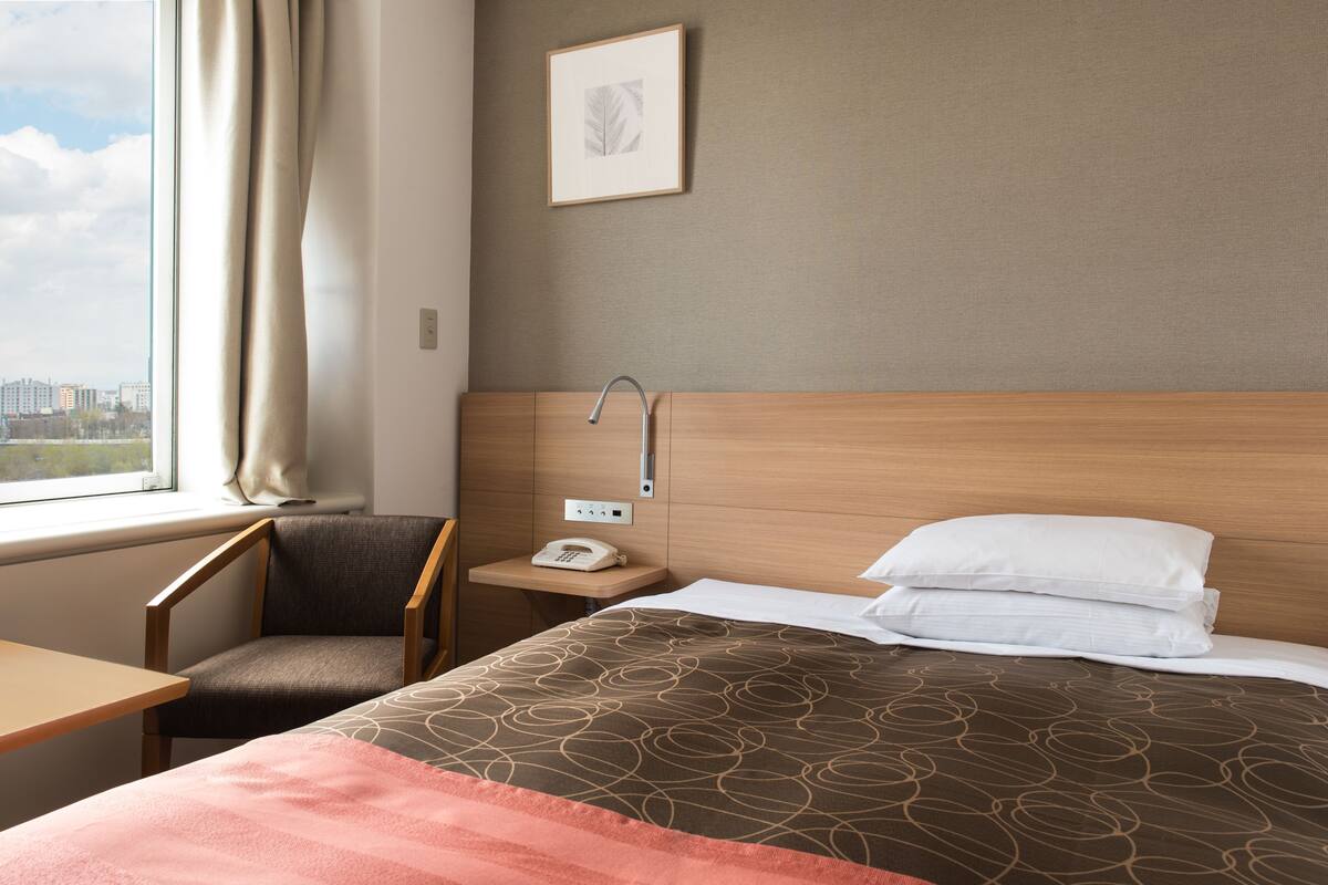 A hotel room with a bed, a chair, a nightstand with a phone, a window with curtains, and a framed picture on the wall.