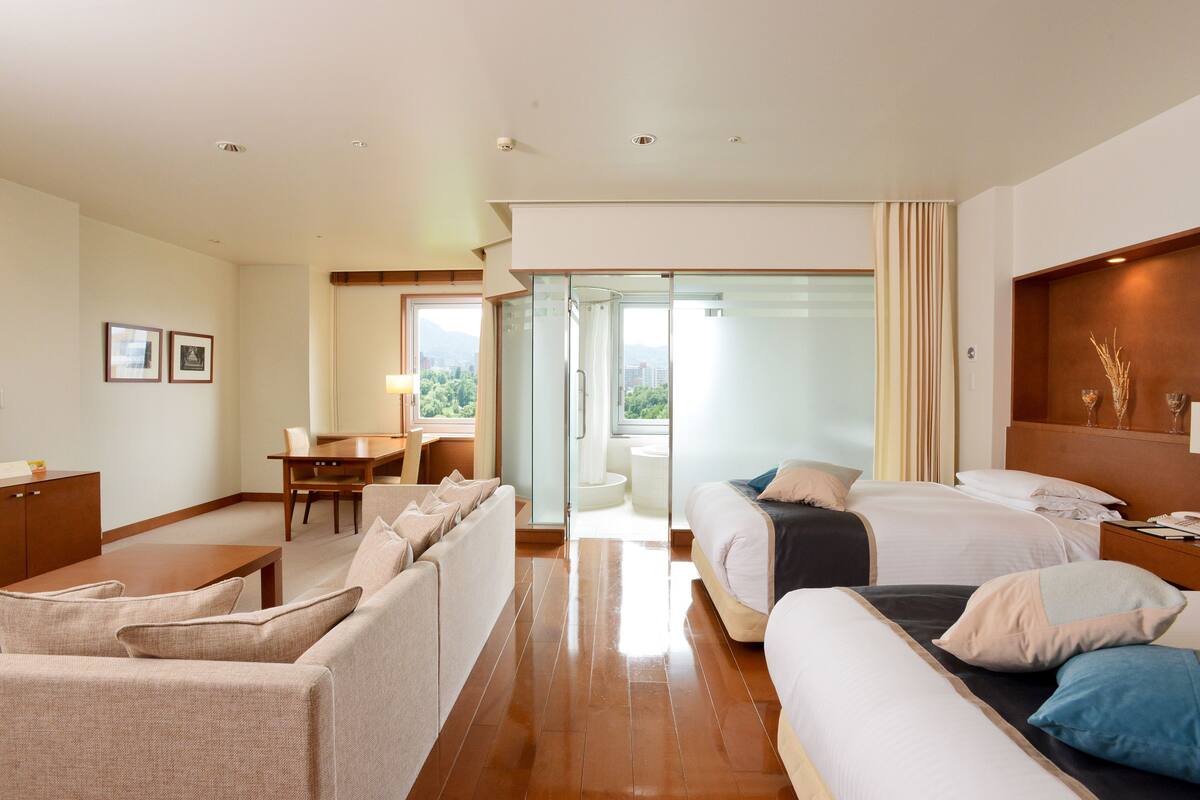 A hotel room with a sofa, two beds, a desk, and a view of the outdoors.