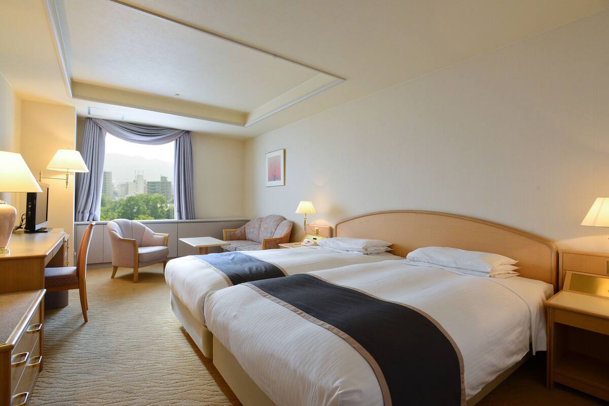 A hotel room with two beds, a desk, a chair, a nightstand, and a window with curtains.