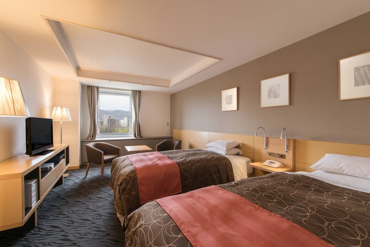 A hotel room with two beds, a desk with a television, a chair, and a window with curtains.