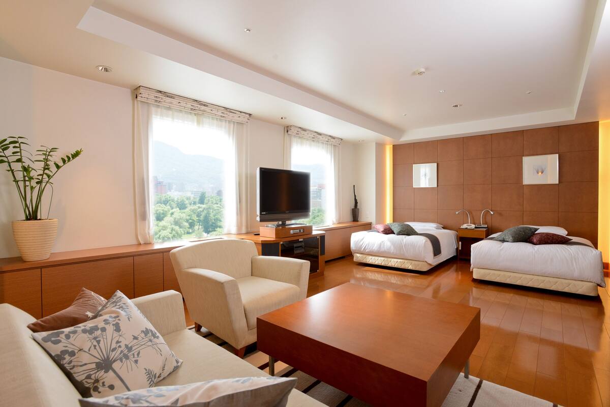 A modern hotel room with a sofa, a coffee table, a TV, two beds, and a large window with a view of the mountains.