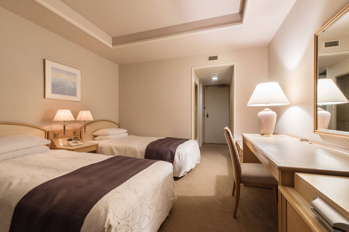 A hotel room with two beds, a desk, a chair, a lamp, a mirror, and a picture on the wall.