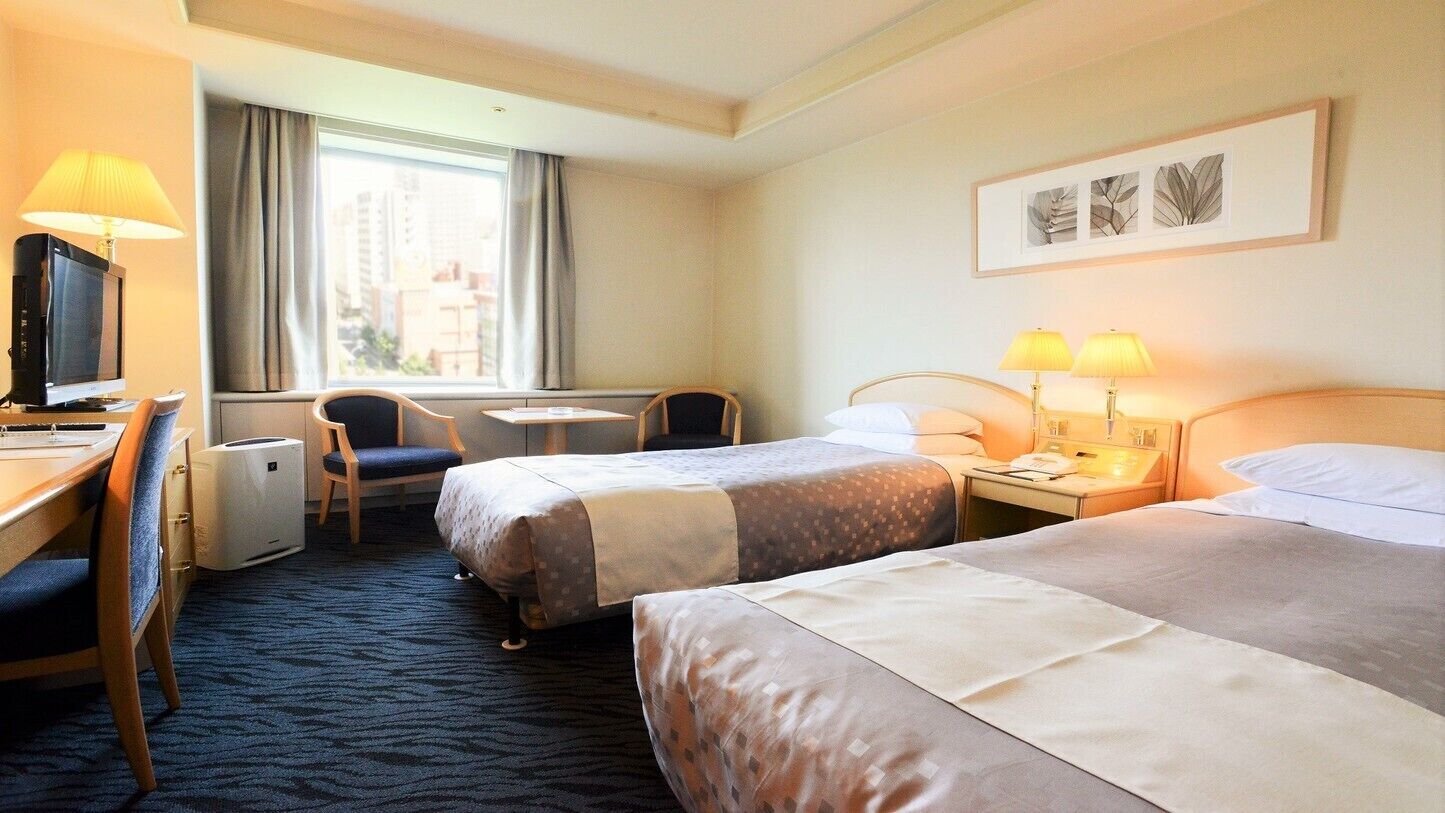 Een hotelkamer met twee bedden, een bureau met een computer, een stoel, een tafeltje en een raam met gordijnen.